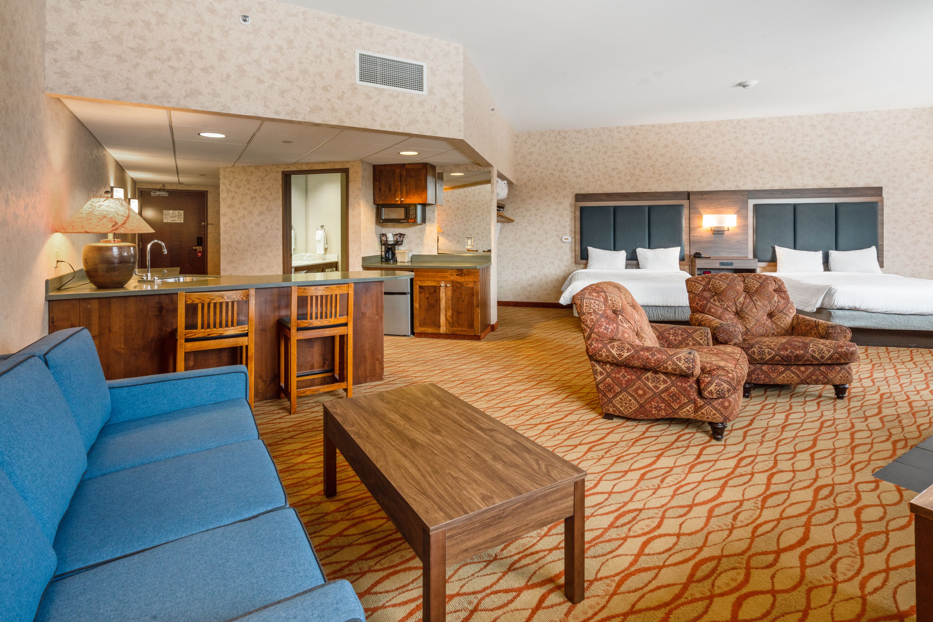 A hotel room with a sofa, two patterned armchairs, a wooden coffee table, a bed with two pillows, a nightstand, a lamp, a microwave, and a small kitchen area.