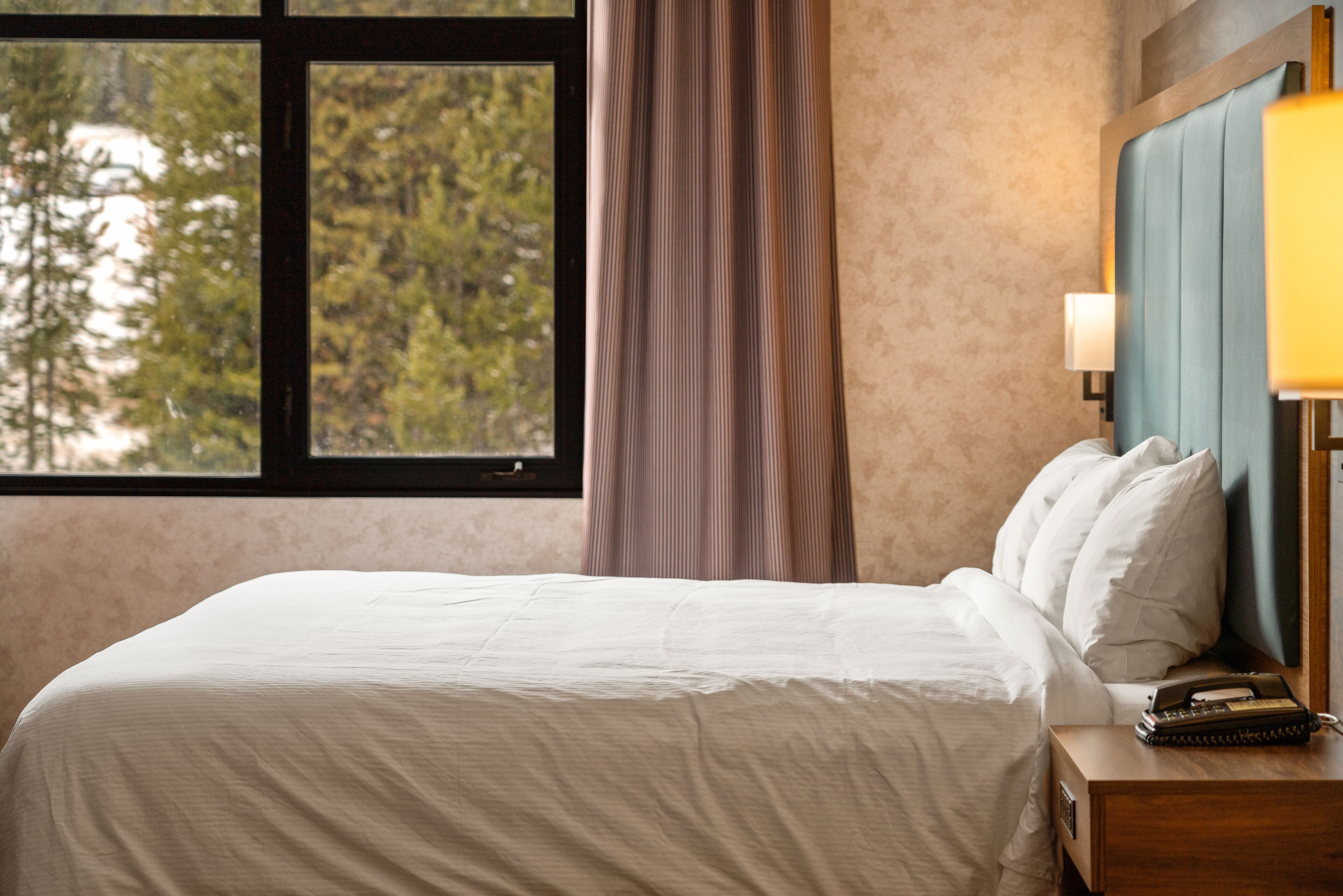 Een hotelkamer met een bed, nachtkastje, lamp en een raam met gordijnen.