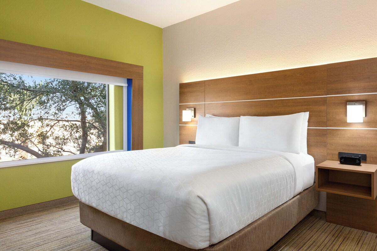 A modern hotel room with a large bed, a nightstand, and a window with a view of trees.