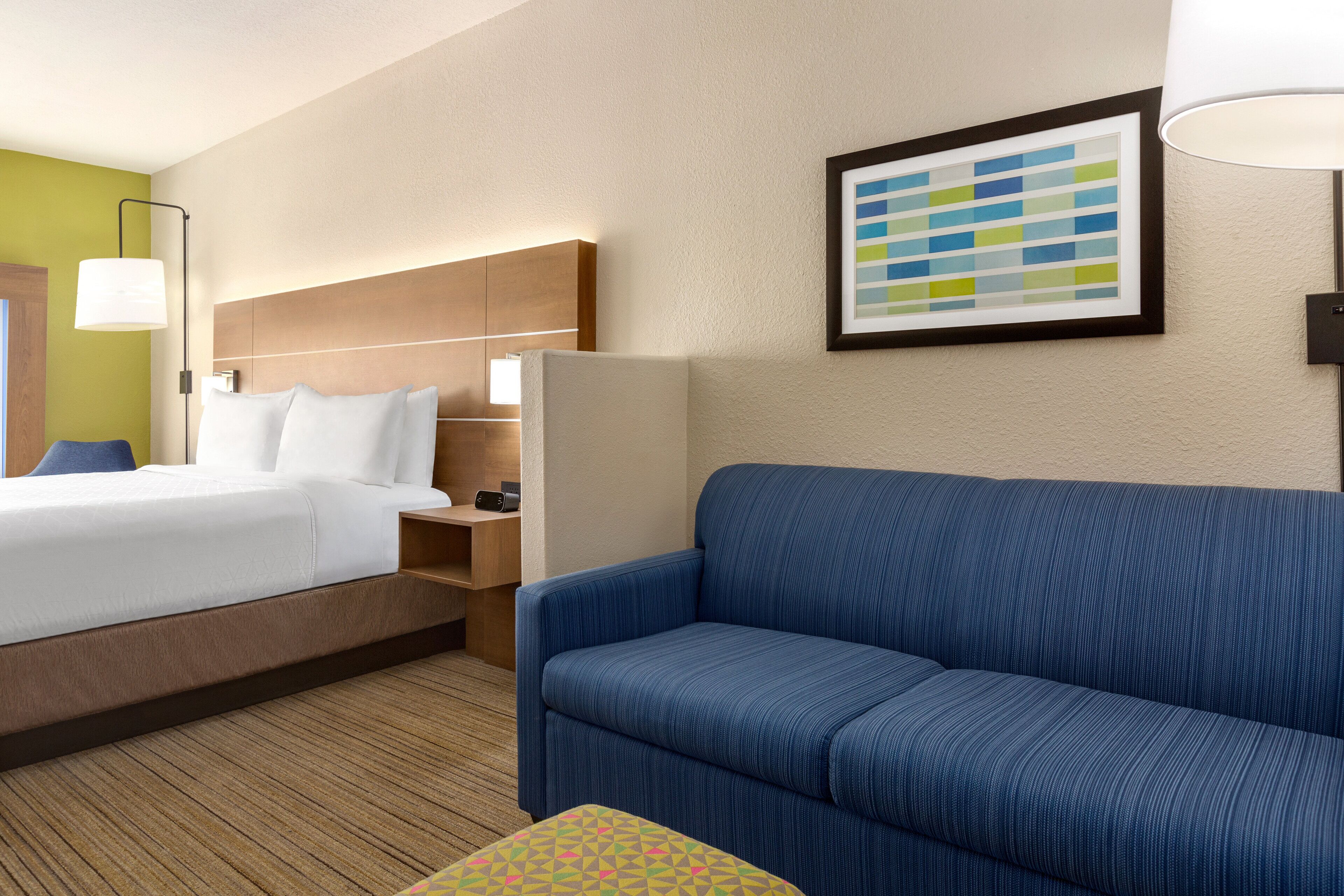 A hotel room with a bed, a blue sofa, a small table, and a wall-mounted lamp.