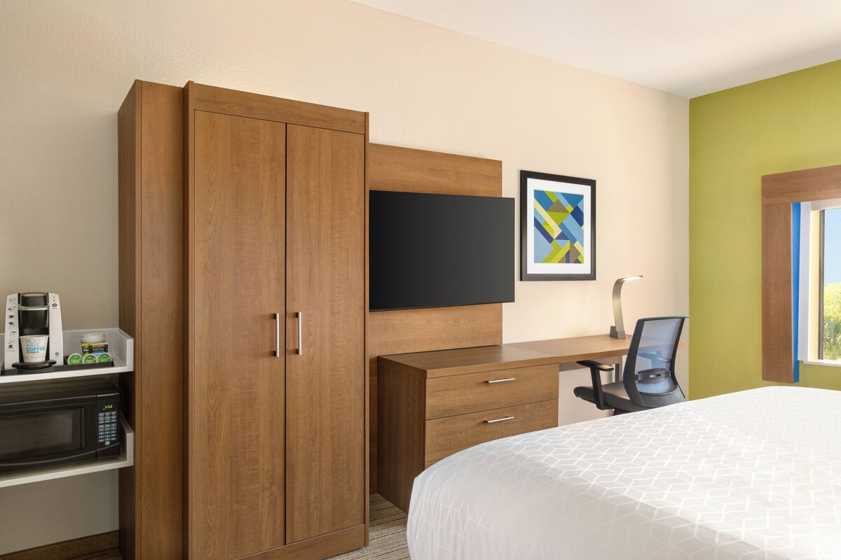 A hotel room with a bed, a wooden wardrobe, a desk with a chair, a flat-screen TV, and a coffee machine.