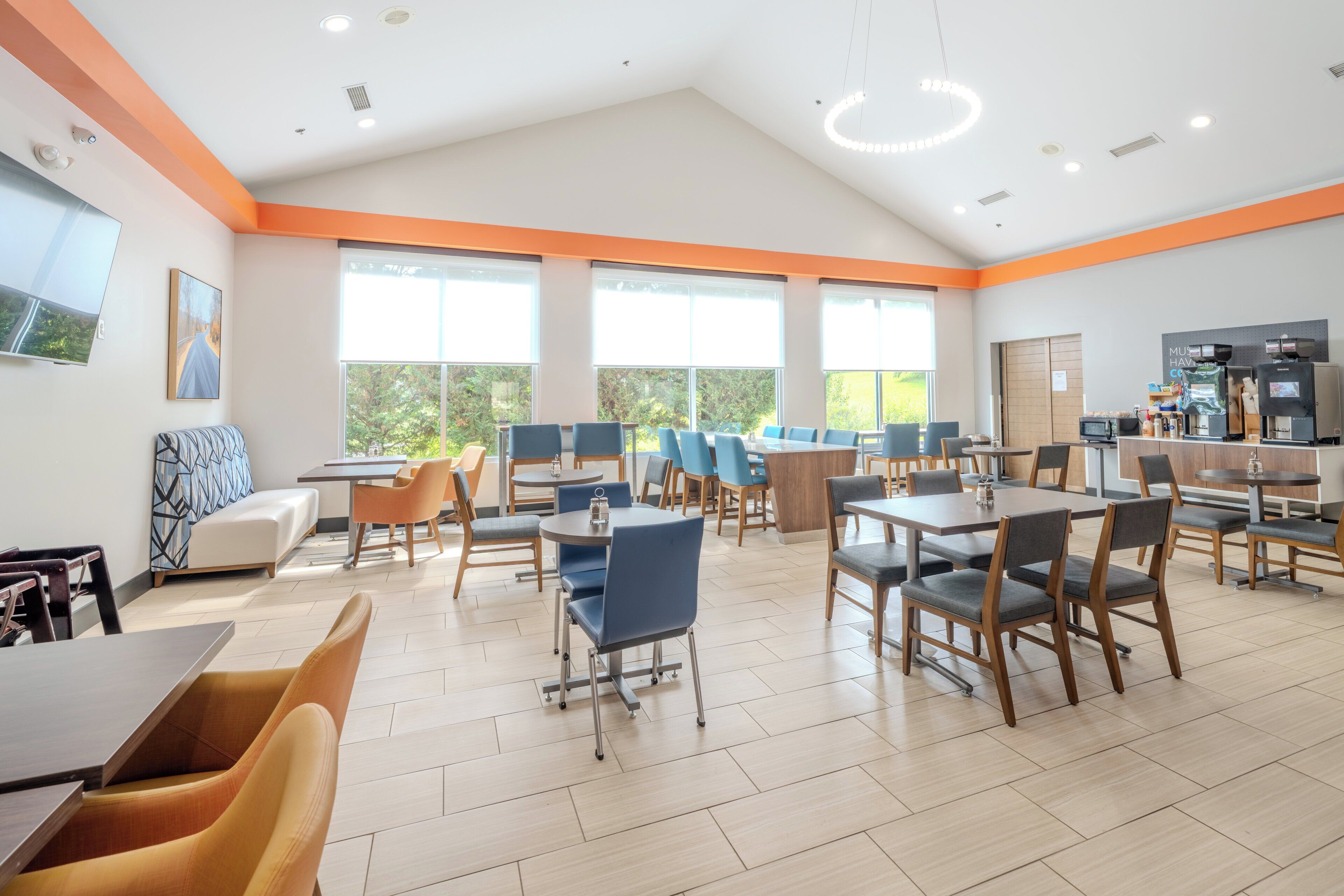 A modern dining area with multiple tables and chairs, large windows, and a kitchen area with appliances.