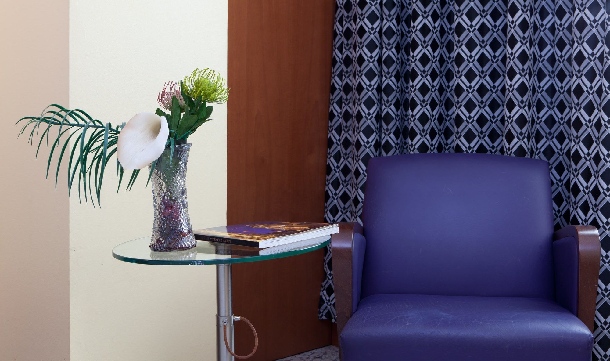 Een kamer met een glazen tafel waarop een vaas met bloemen, een boek en een stoel staan. Op de achtergrond een gordijn met een patroon.