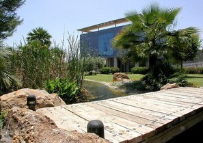 A modern building with a glass facade, surrounded by a landscaped garden with a wooden deck, palm trees, and a small pond. - Hotel Jardines de Amaltea (Lorca)