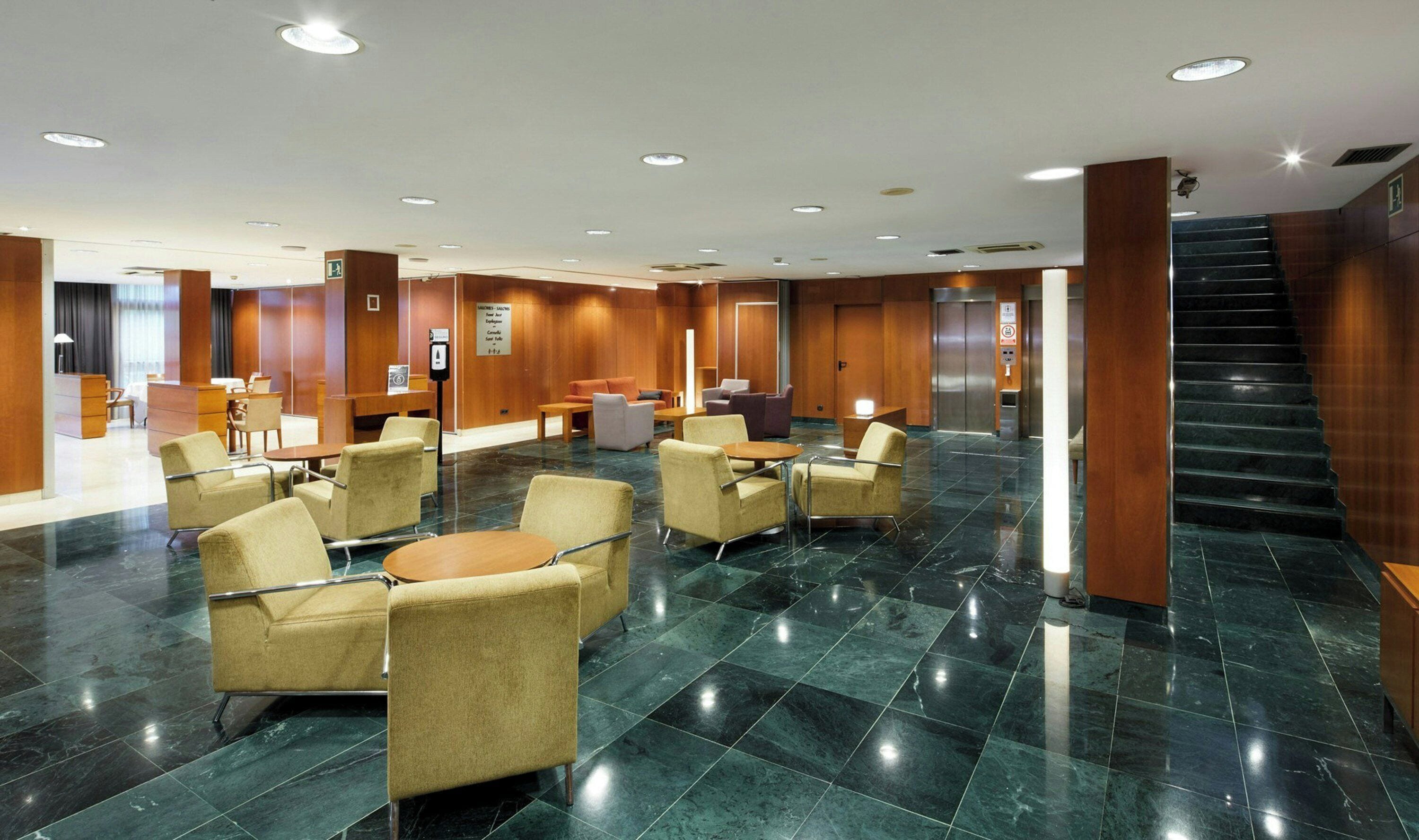 Um hall amplo com área de estar central, balcão de recepção e escada que dá acesso a um elevador.