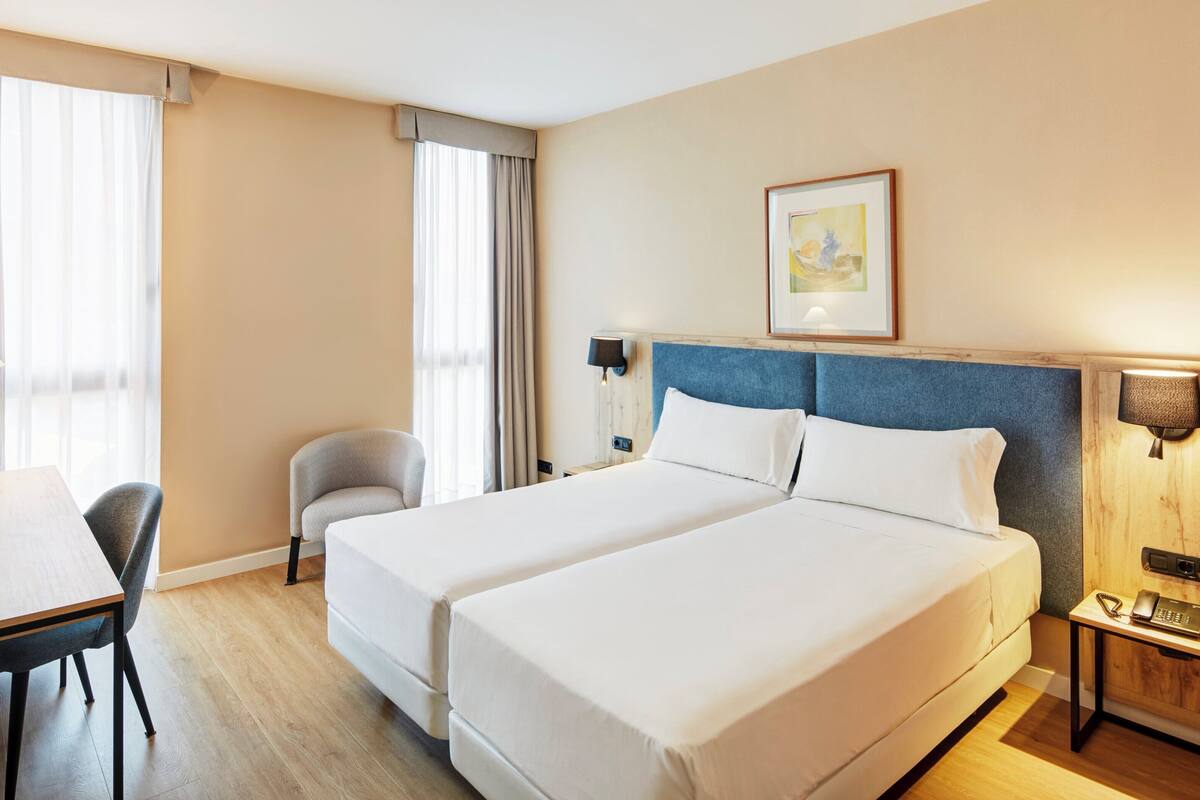 A hotel room with two beds, a desk, a chair, a lamp, and a painting on the wall.