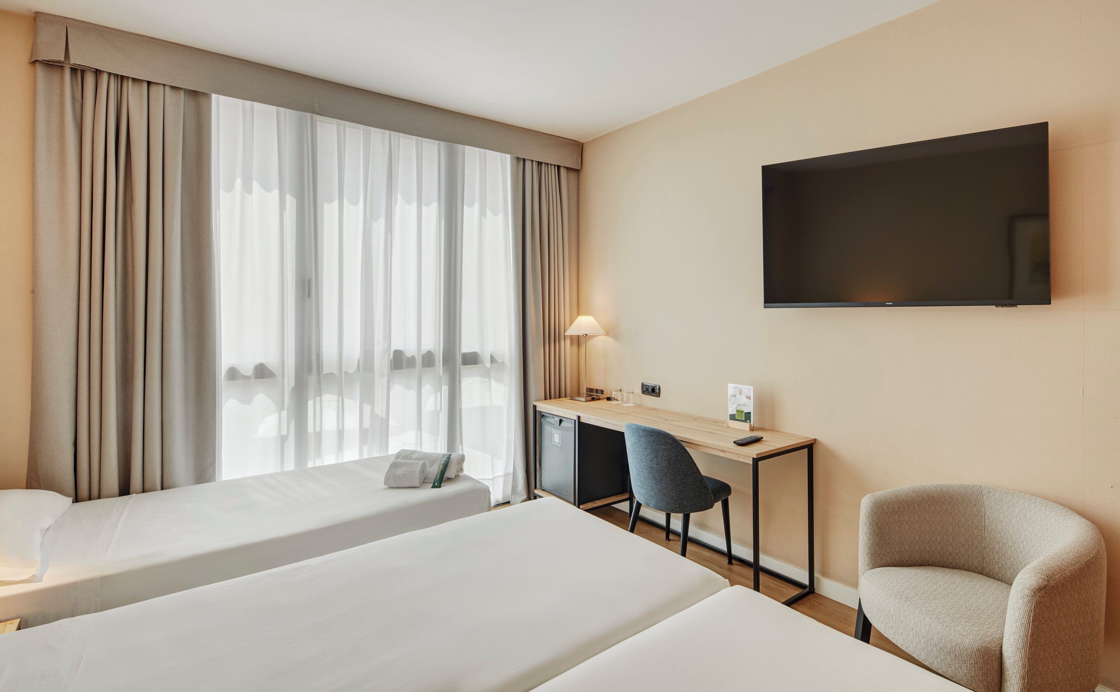 A hotel room with two beds, a desk, a chair, a TV, and a window with curtains.