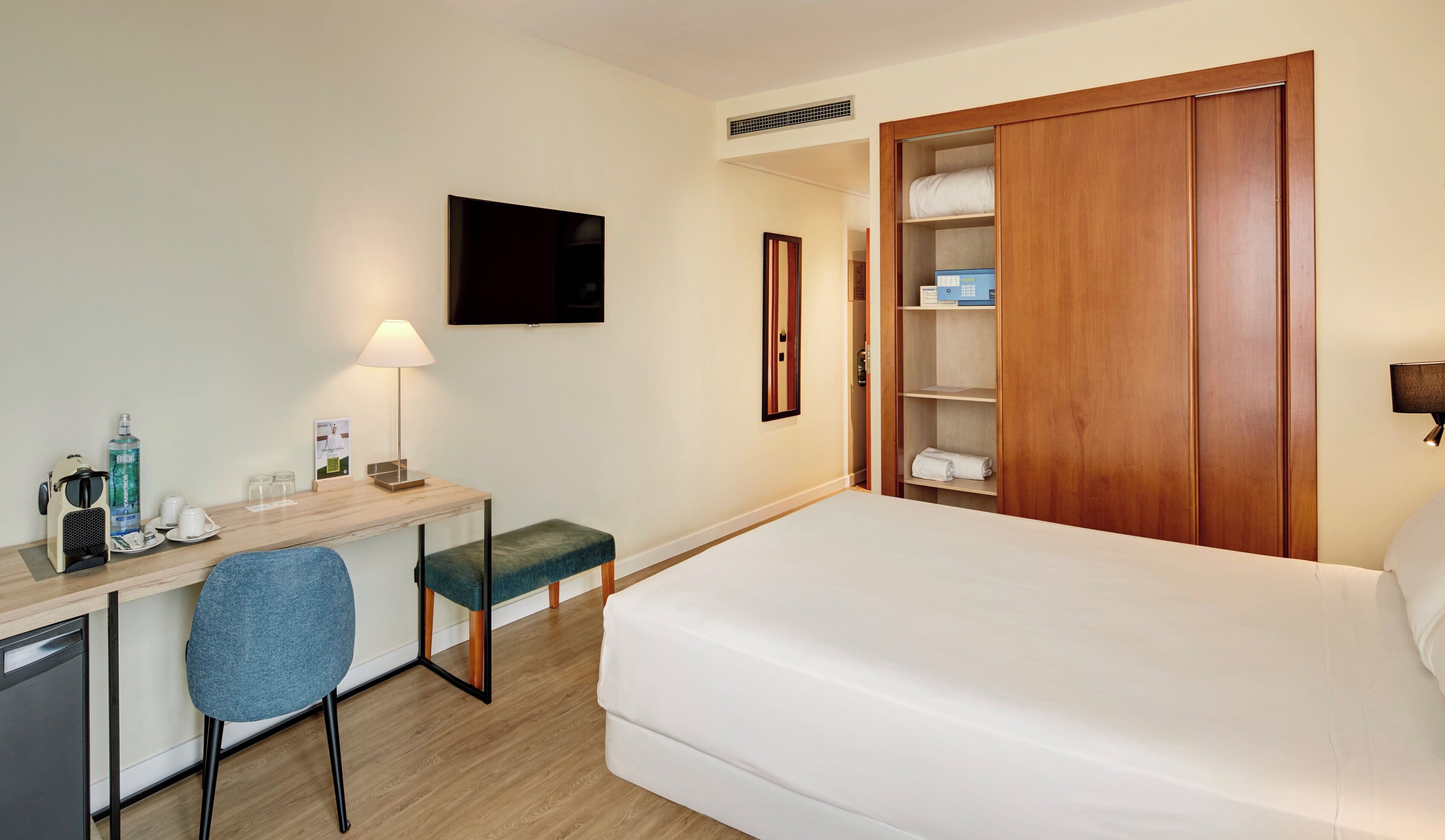 Habitación de hotel con cama, escritorio y silla, televisión, mesita de noche con lámpara y armario.