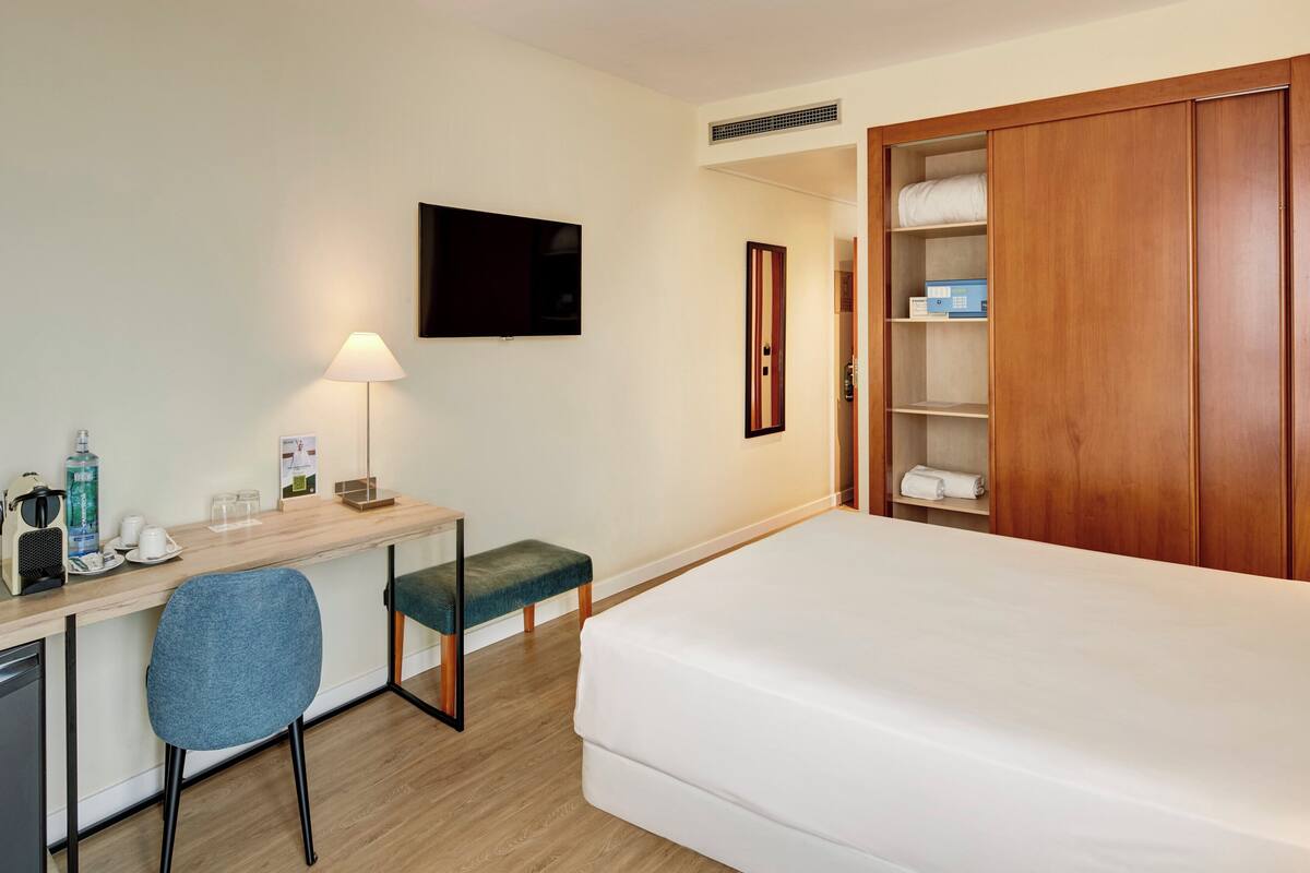 A hotel room with a bed, a desk with a chair, a TV, a bedside table with a lamp, and a wardrobe.