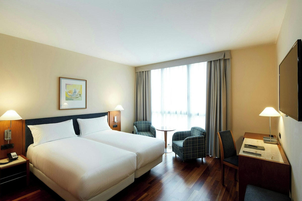 A hotel room with a double bed, two bedside lamps, a desk with a chair, a small table, and a large window with curtains.