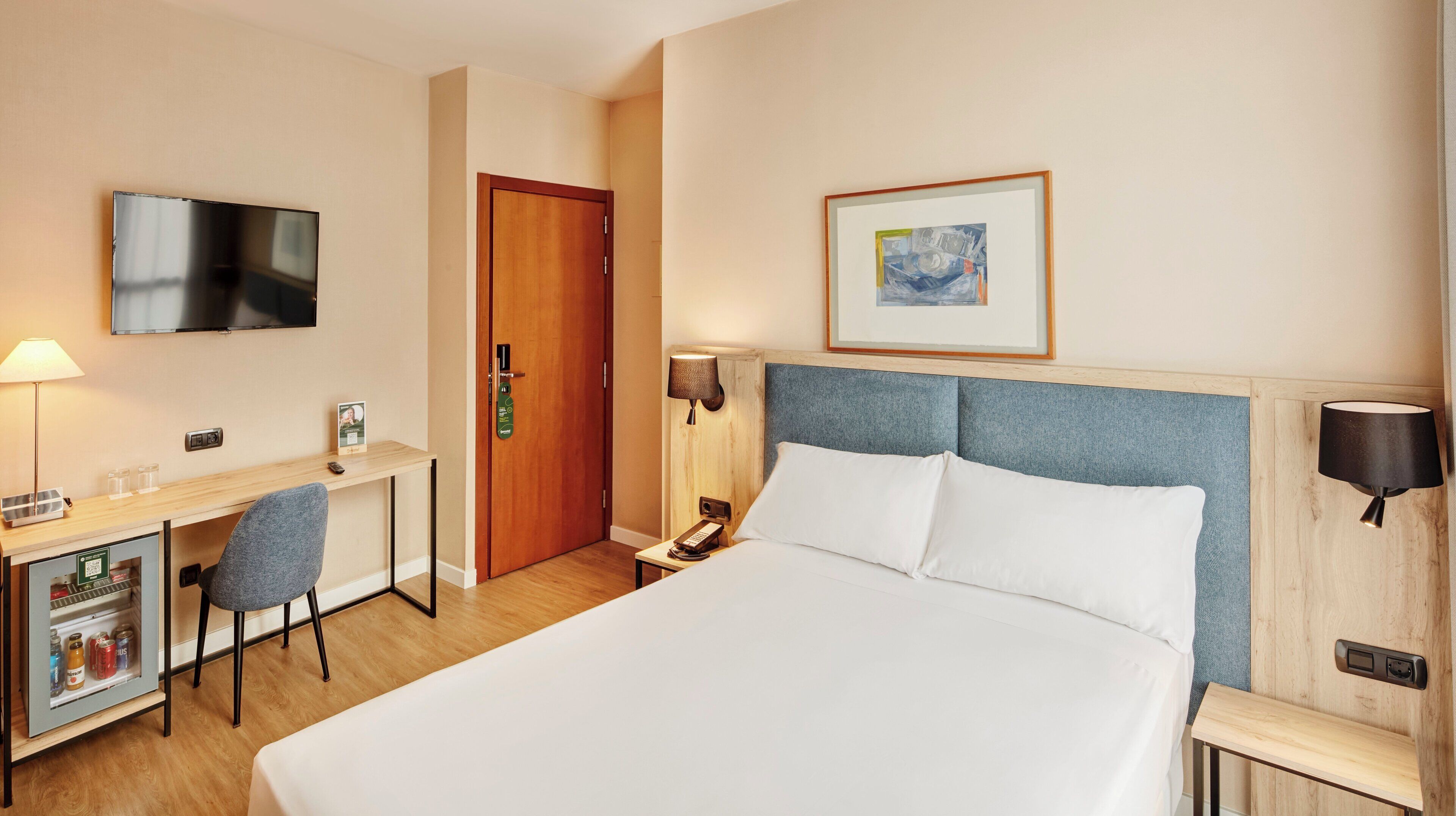 Habitación de hotel con cama, escritorio, silla, televisor, puerta, un cuadro enmarcado y mesita de noche con lámpara.