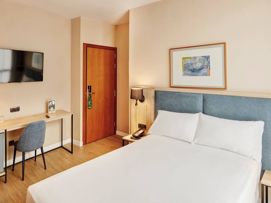 Habitación de hotel con cama, escritorio, silla, televisor, puerta, un cuadro enmarcado y mesita de noche con lámpara.