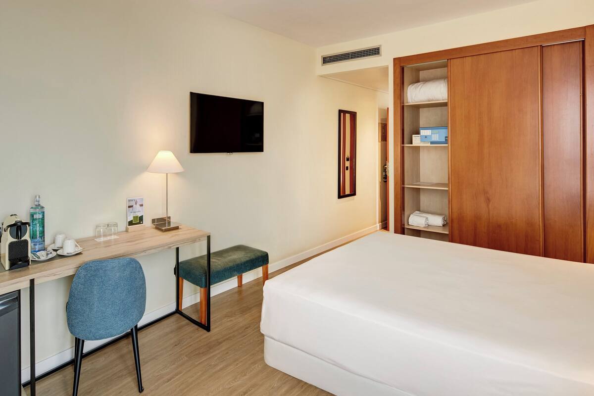 Habitació d'hotel amb un llit, una escriptori amb una cadira, una televisió, una taula de nit amb una làmpada i un armari.