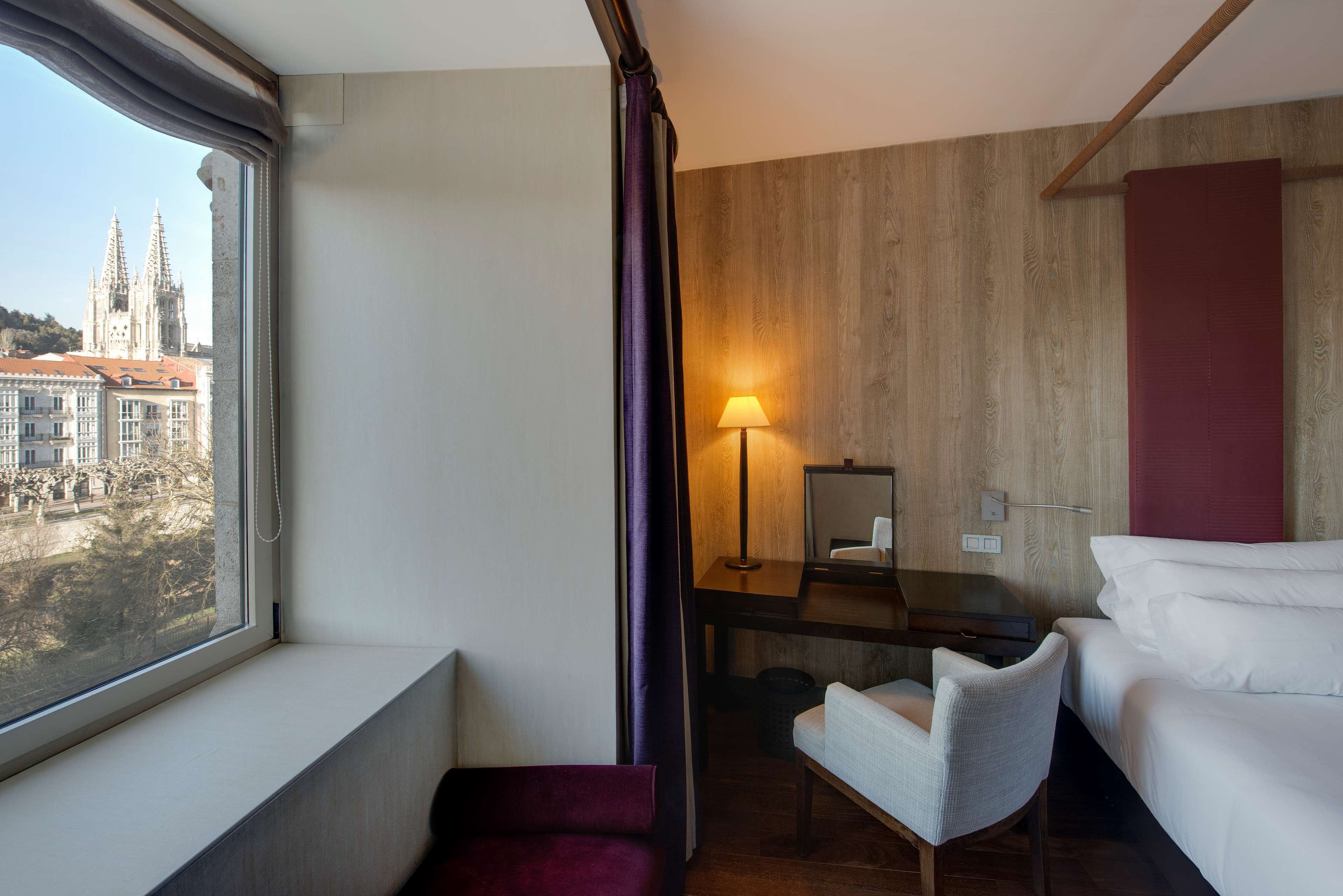 A hotel room with a large window, a desk with a lamp, a chair, and a bed.