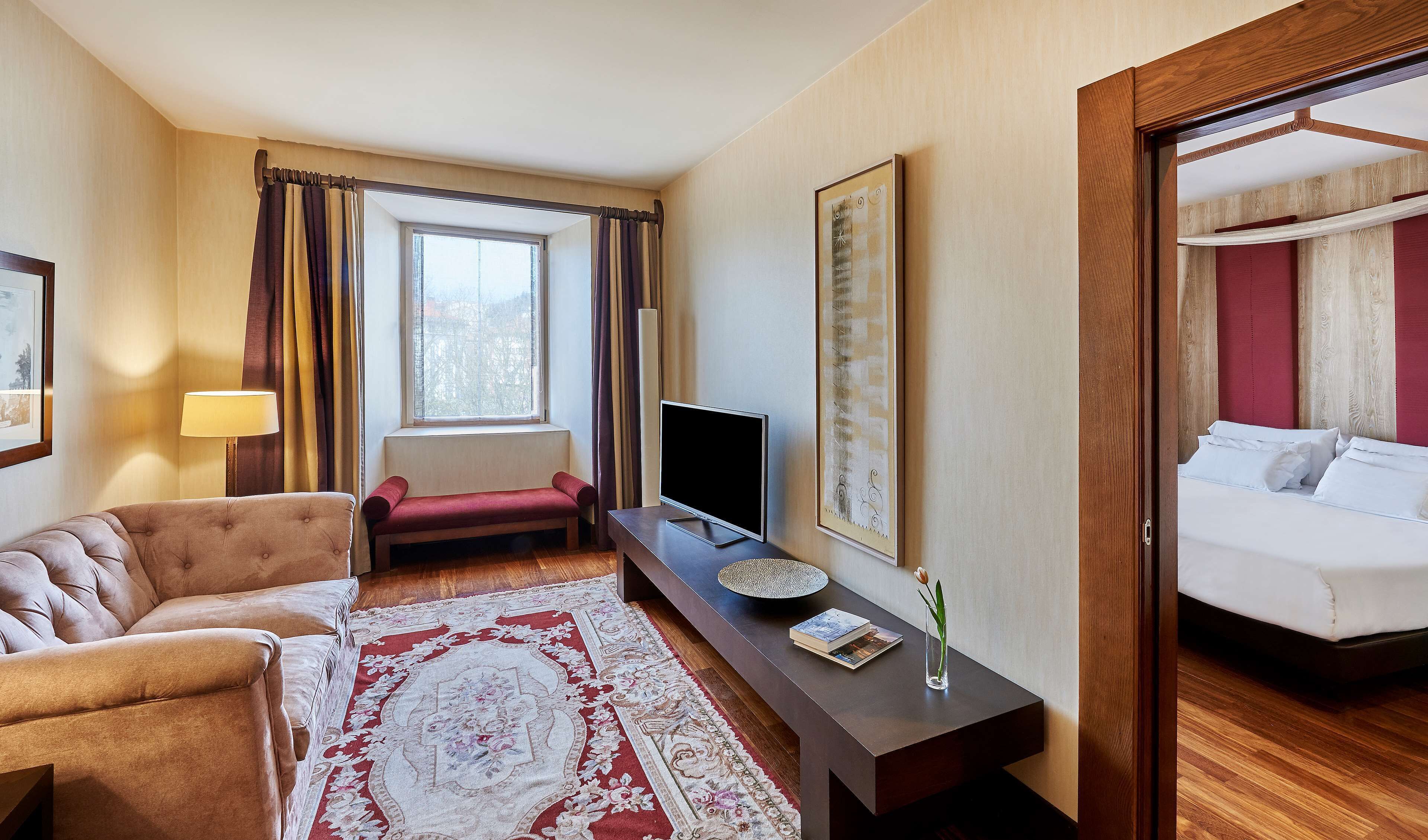 Habitación de hotel con sofá, televisor en un mueble bajo, cama, mesita de noche con un libro y un florero, y ventana con cortinas.