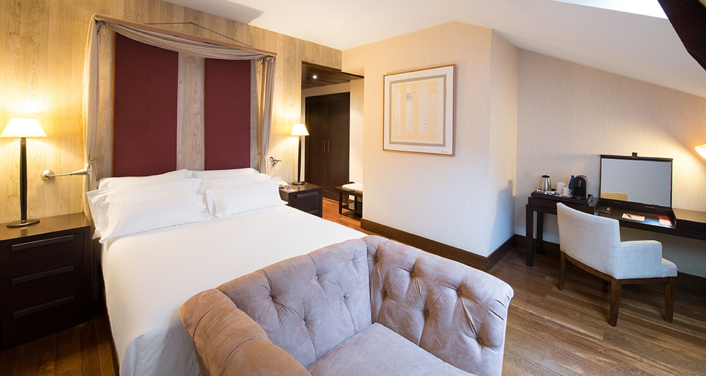 Une chambre d’hôtel avec un grand lit, un coin salon avec un fauteuil, un bureau avec un ordinateur et un tableau encadré au mur.