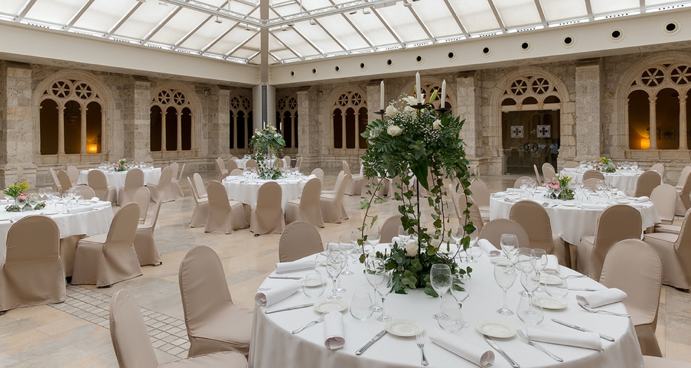 A banquet hall with round tables set for a formal event, featuring white tablecloths, floral centerpieces, and beige chairs.
