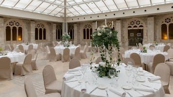 A banquet hall with round tables set for a formal event, featuring white tablecloths, floral centerpieces, and beige chairs.