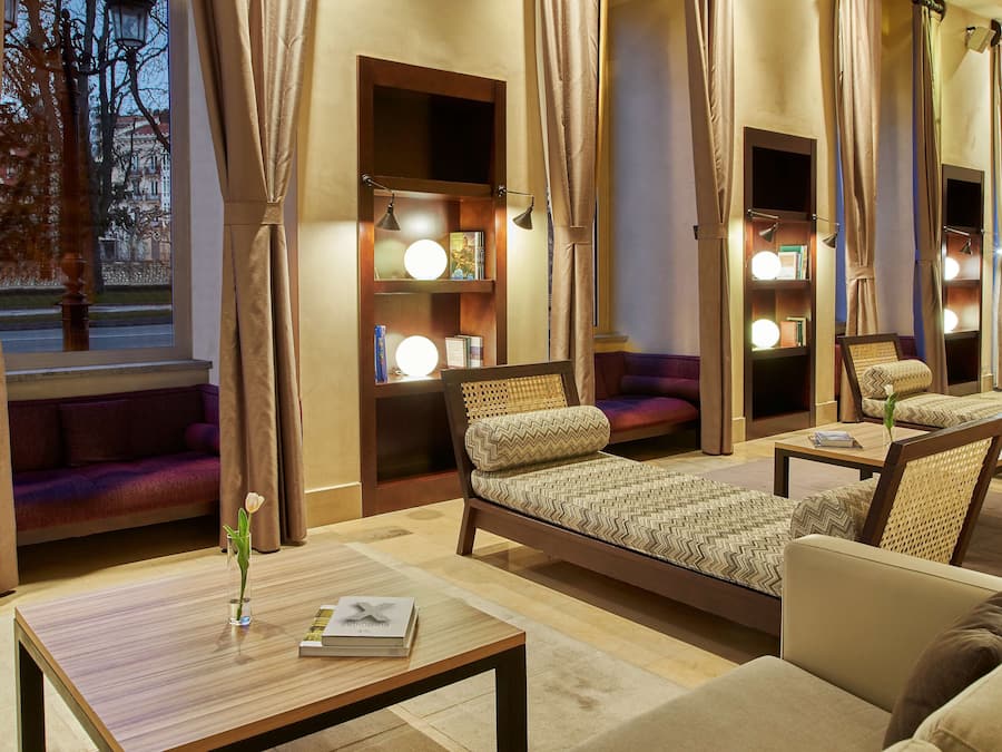 Habitación luminosa con cama, sofá y mesa. Hay cortinas, una lámpara y una ventana con vistas a edificios.