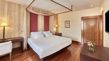 A hotel room with a large bed, wooden flooring, a desk with a lamp, a bedside table with a lamp, and a door.