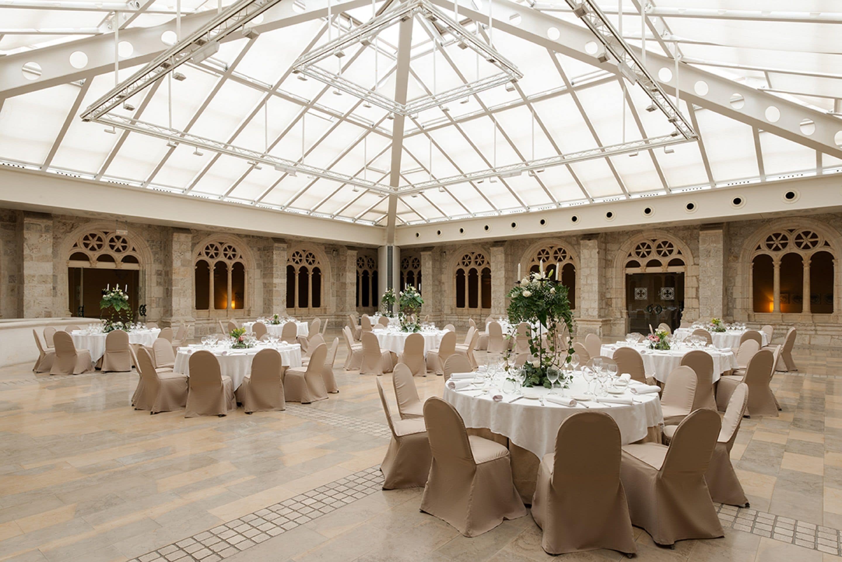 A spacious hall with a high ceiling and a glass roof, featuring multiple round tables set for dining, surrounded by beige chairs.