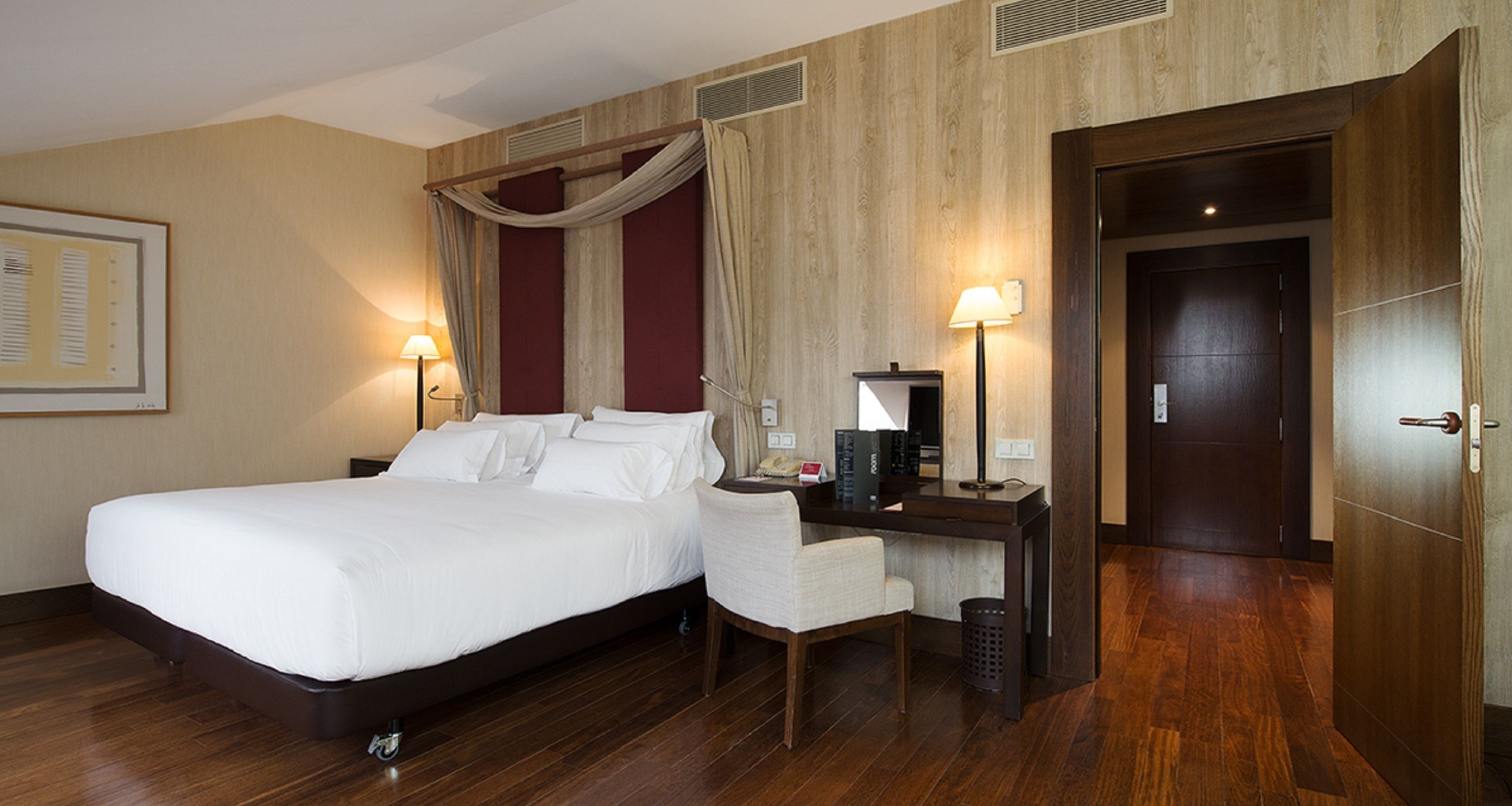 Habitación de hotel con una cama grande, un escritorio con silla, una lámpara y una puerta.