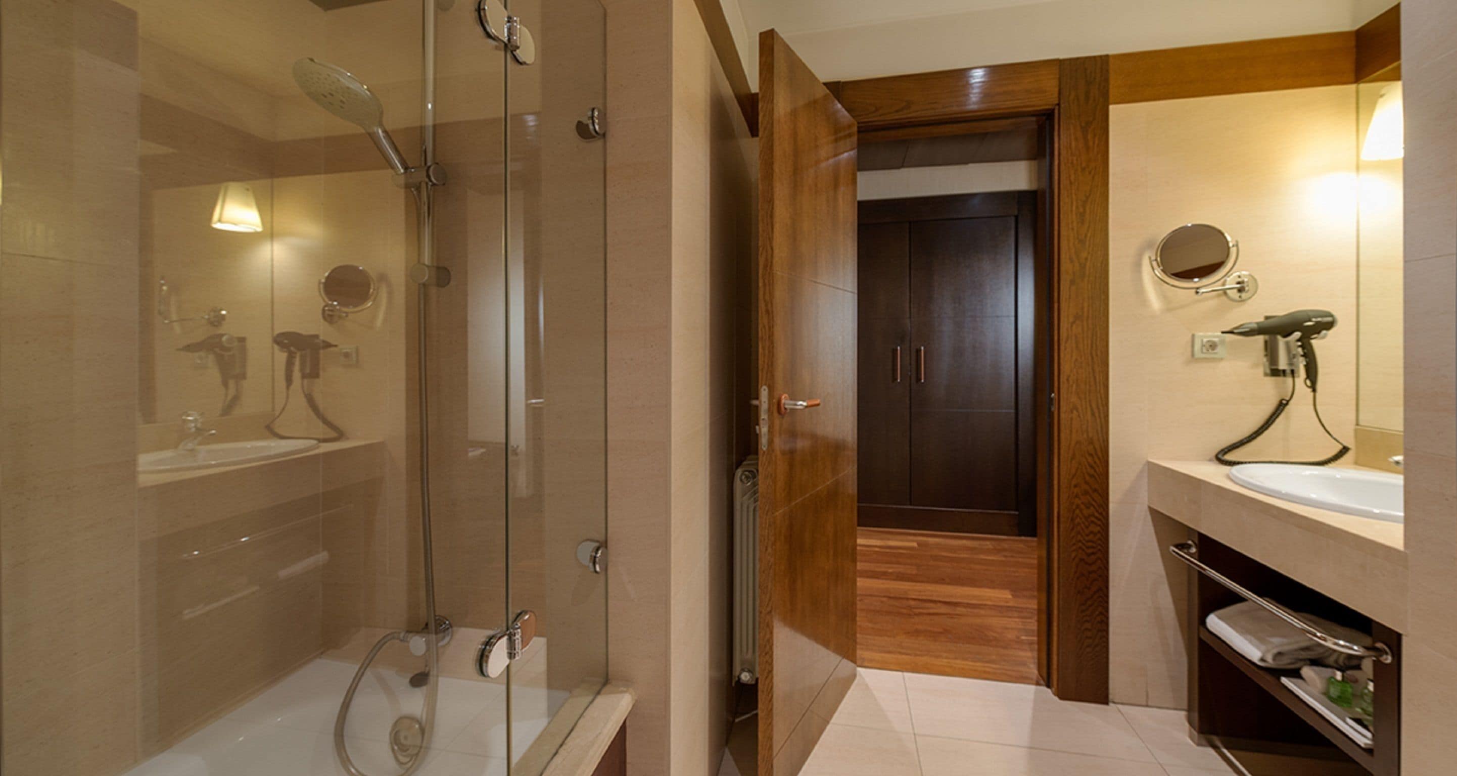 A modern bathroom with a glass-enclosed shower, a wooden door leading to a separate room, and a vanity with a mirror and hairdryer.
