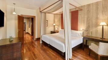 Quarto de hotel com cama de dossel, piso de madeira, mesa de cabeceira com abajur e uma pequena mesa com um vaso de flores.