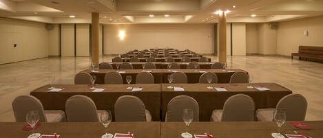 Un salón de conferencias con filas de mesas, cada una dispuesta con sillas y copas de vino, en un espacio amplio y bien iluminado con una paleta de colores neutros.