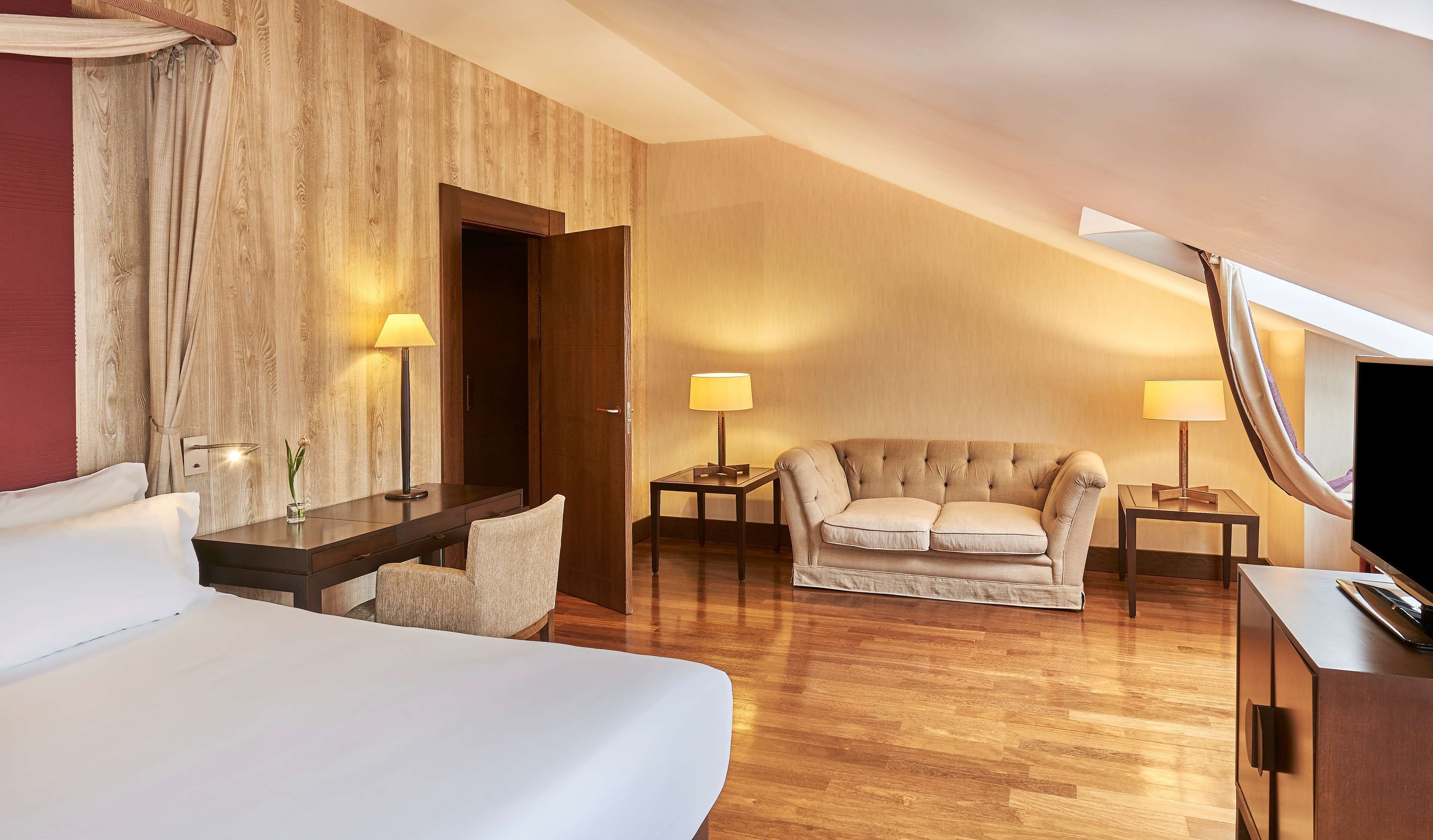 Habitación de hotel con una cama grande, un sofá, un escritorio y un televisor. Hay lámparas en el escritorio y el sofá, y una pared de madera.