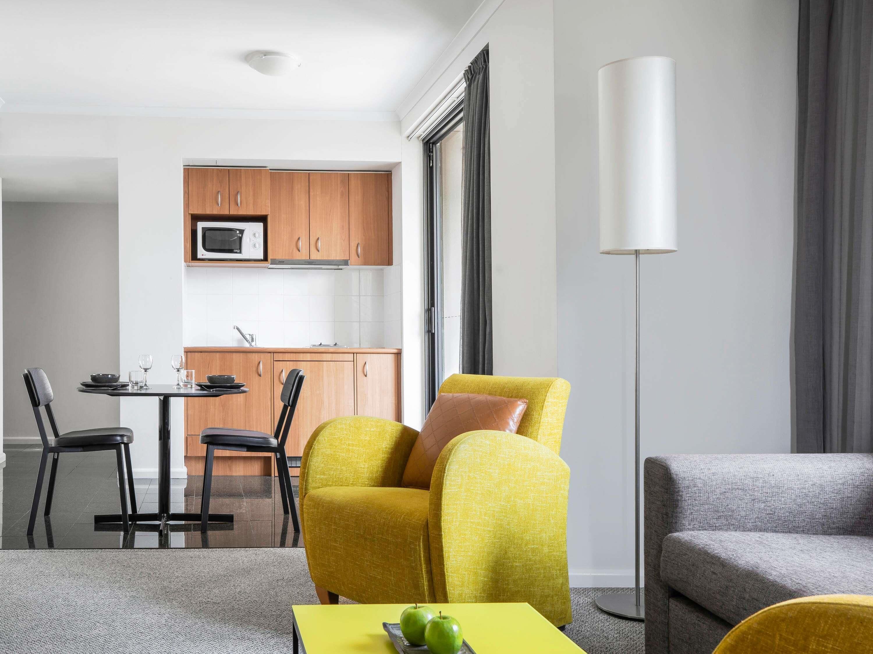 Una sala de estar moderna con un sillón amarillo, un sofá gris, una mesa de comedor con sillas y una zona de cocina al fondo.