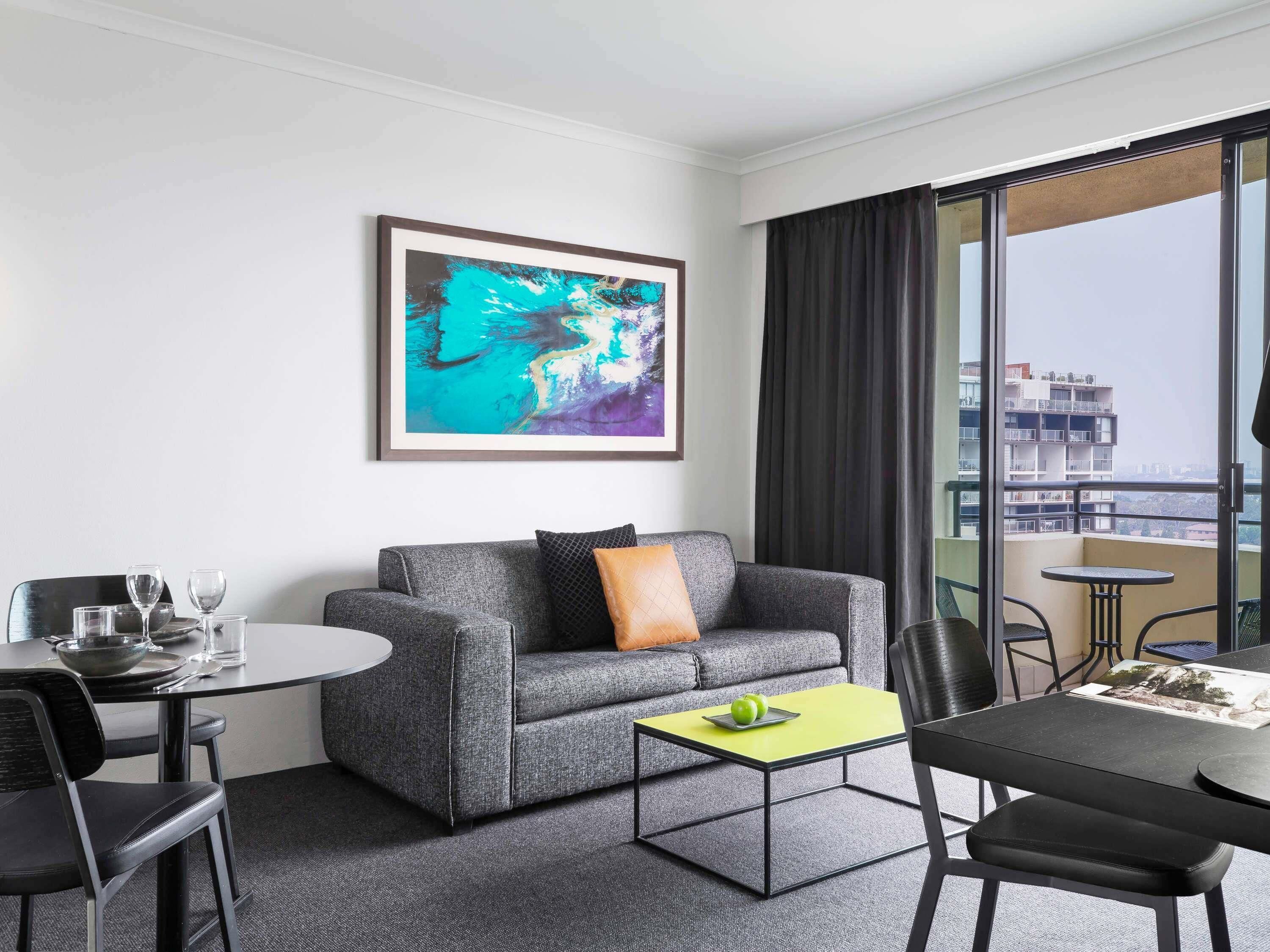 Quarto de hotel com um sofá cinzento, uma mesa redonda pequena, uma mesa de jantar e uma grande janela com vista para edifícios.