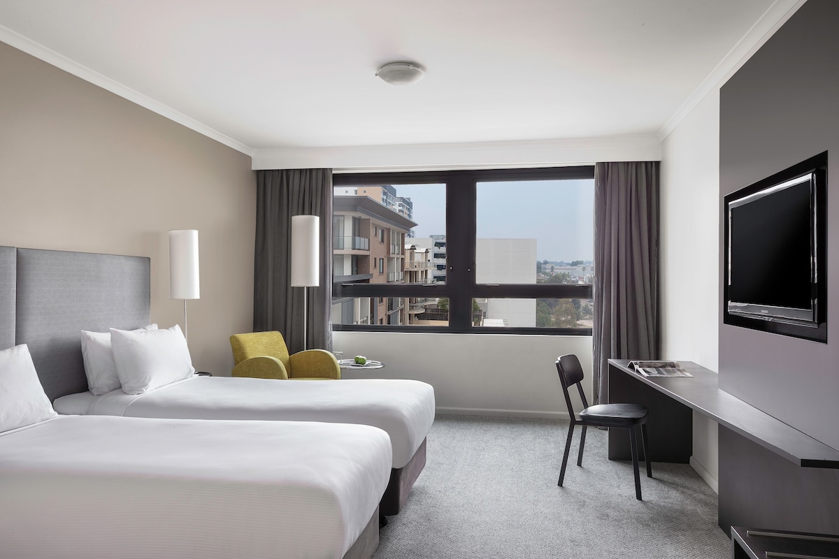 A hotel room with two beds, a desk, a chair, a TV, and a window with a view of buildings.