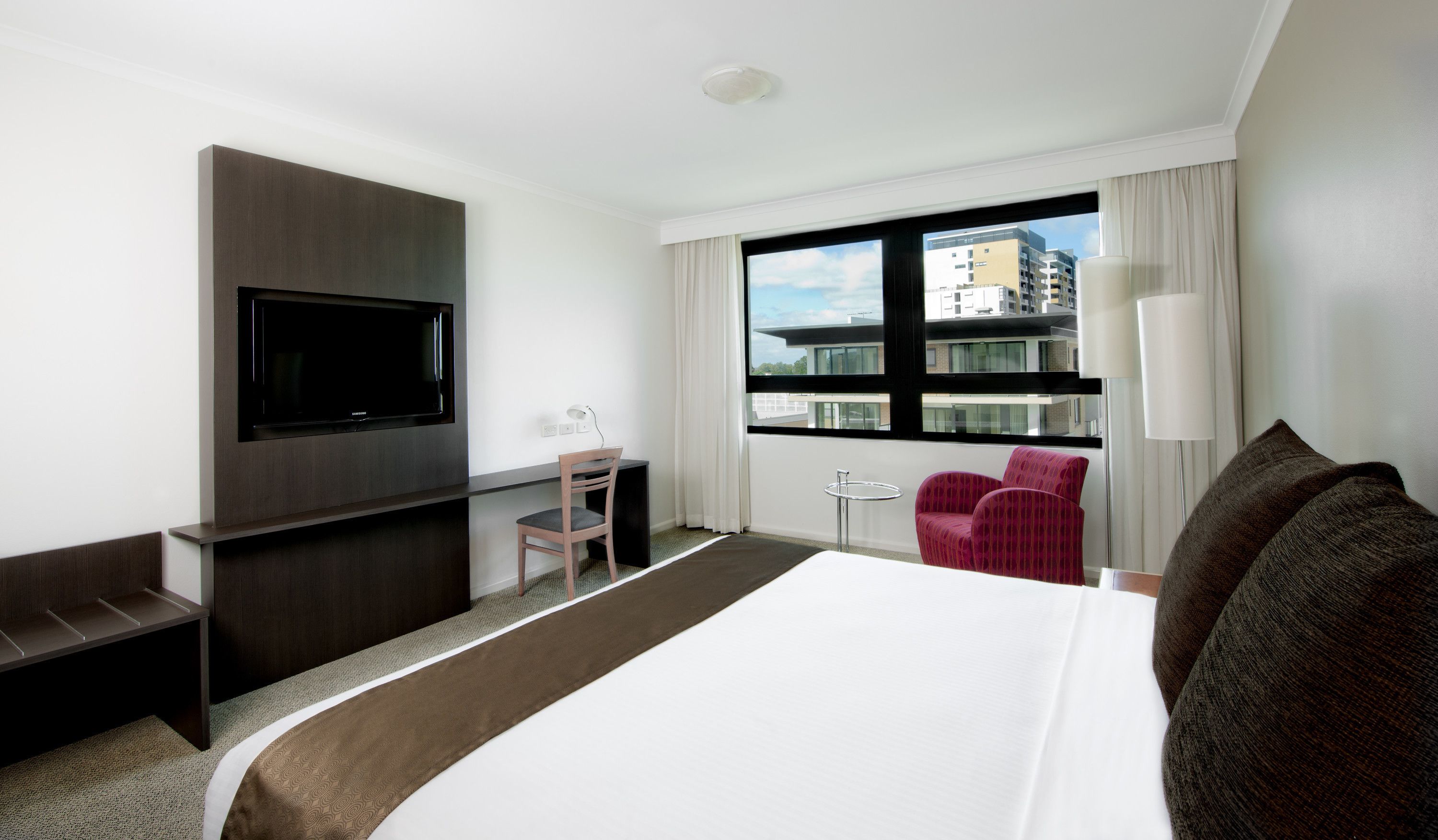 A hotel room with a large bed, a flat-screen TV mounted on the wall, a desk with a chair, a small table, a red chair, and a window with a view of buildings.