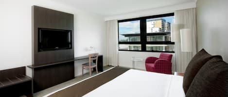 A hotel room with a large bed, a flat-screen TV mounted on the wall, a desk with a chair, a small table, a red chair, and a window with a view of buildings.