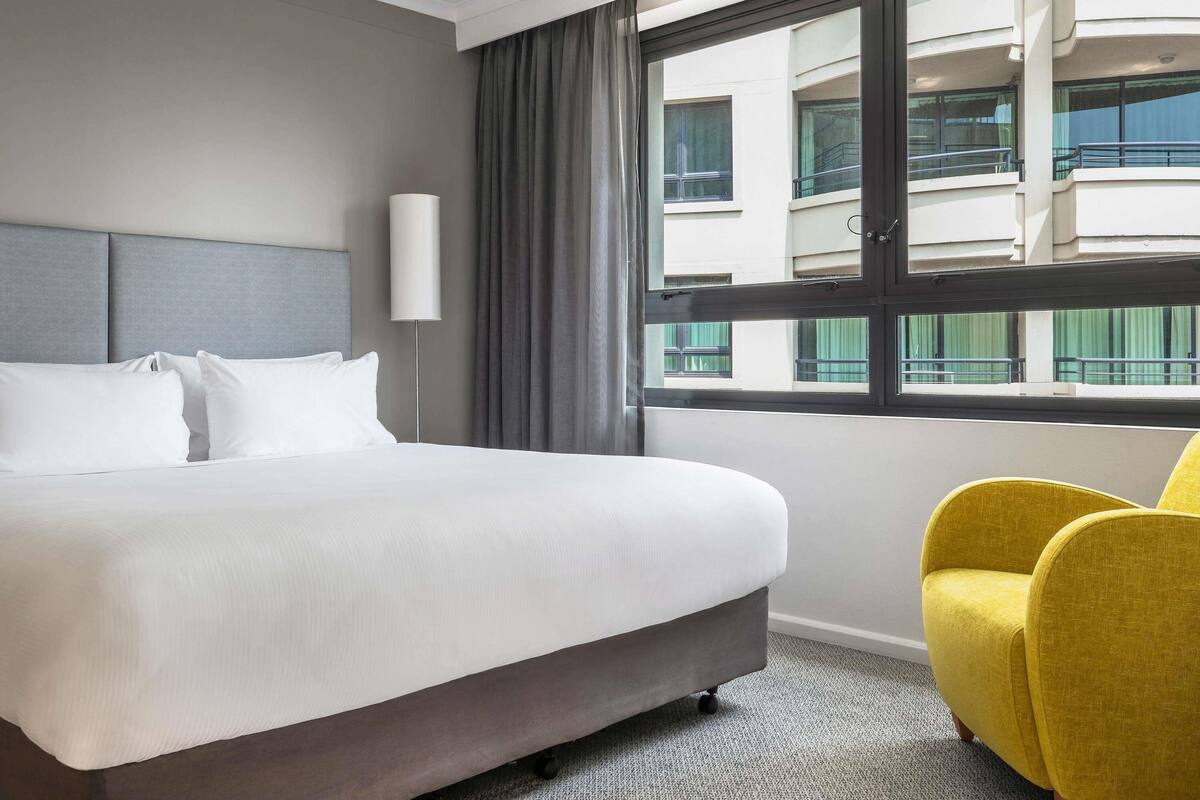 A hotel room with a bed, a chair, a window with a view of buildings, and a wall-mounted light.