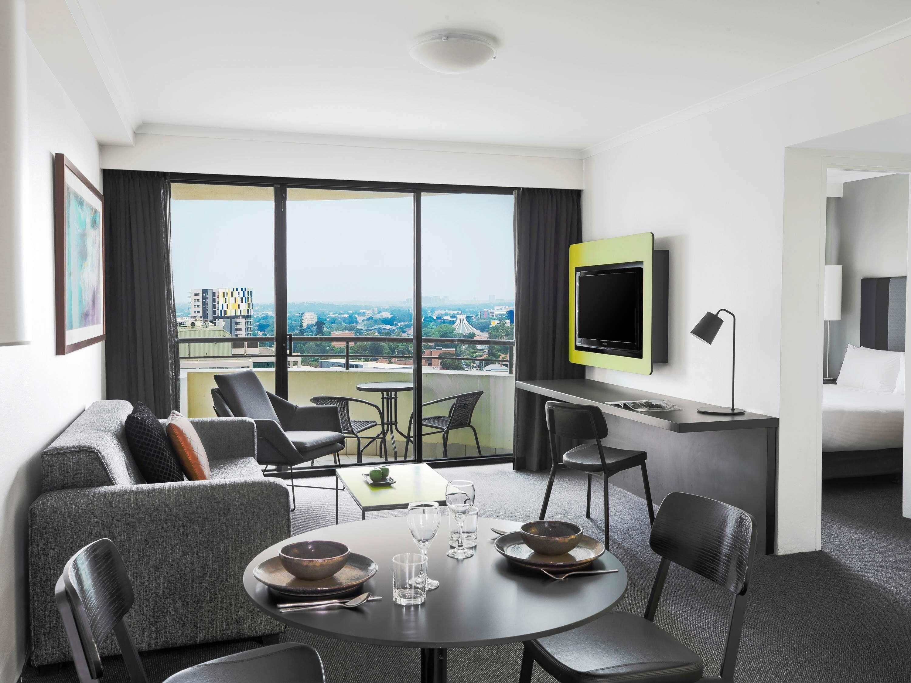 A modern hotel room with a balcony, a flat-screen TV, a dining area, and a bed.