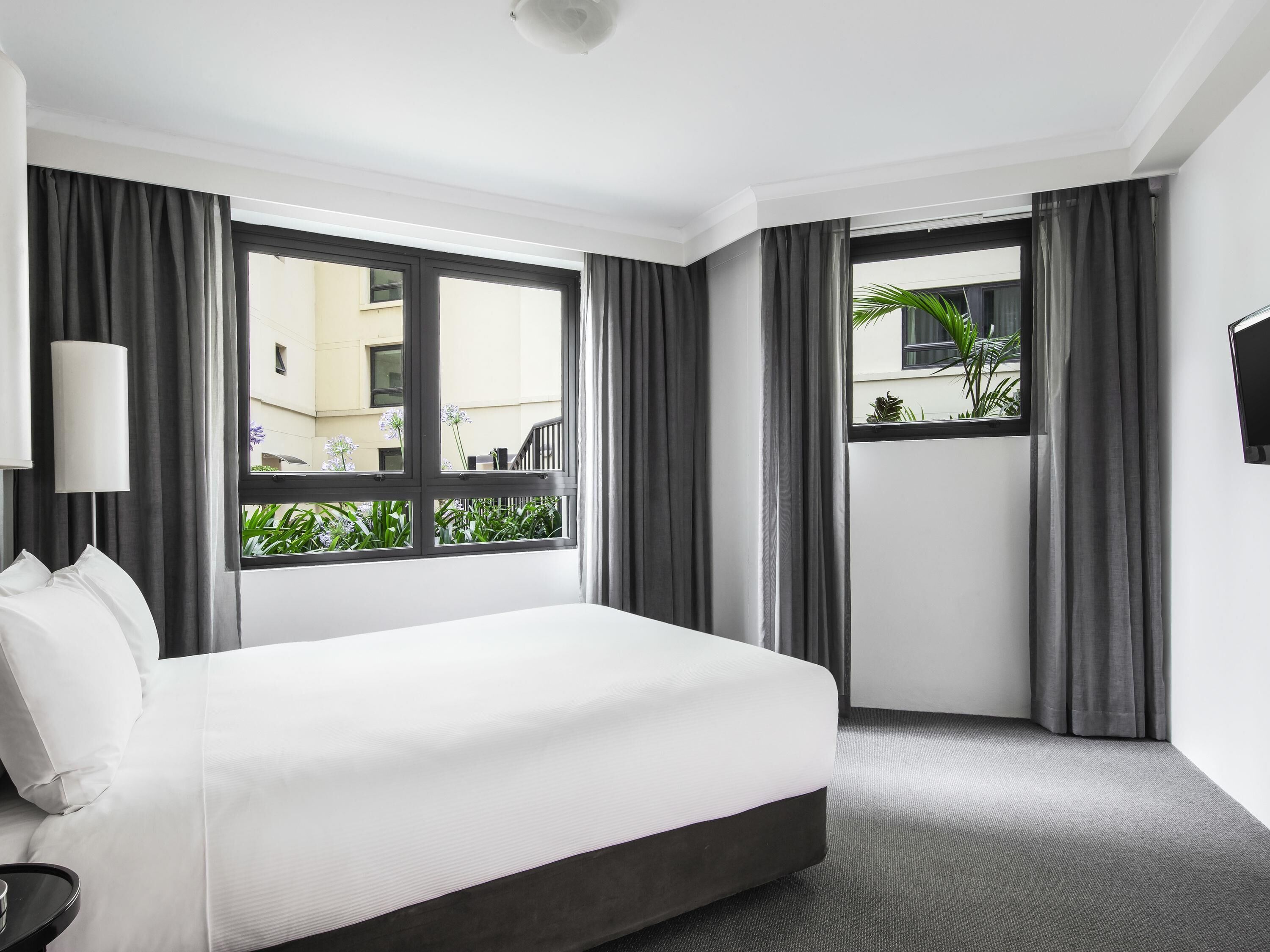 A hotel room with a bed, a TV, a window with curtains, and a view of the outside.