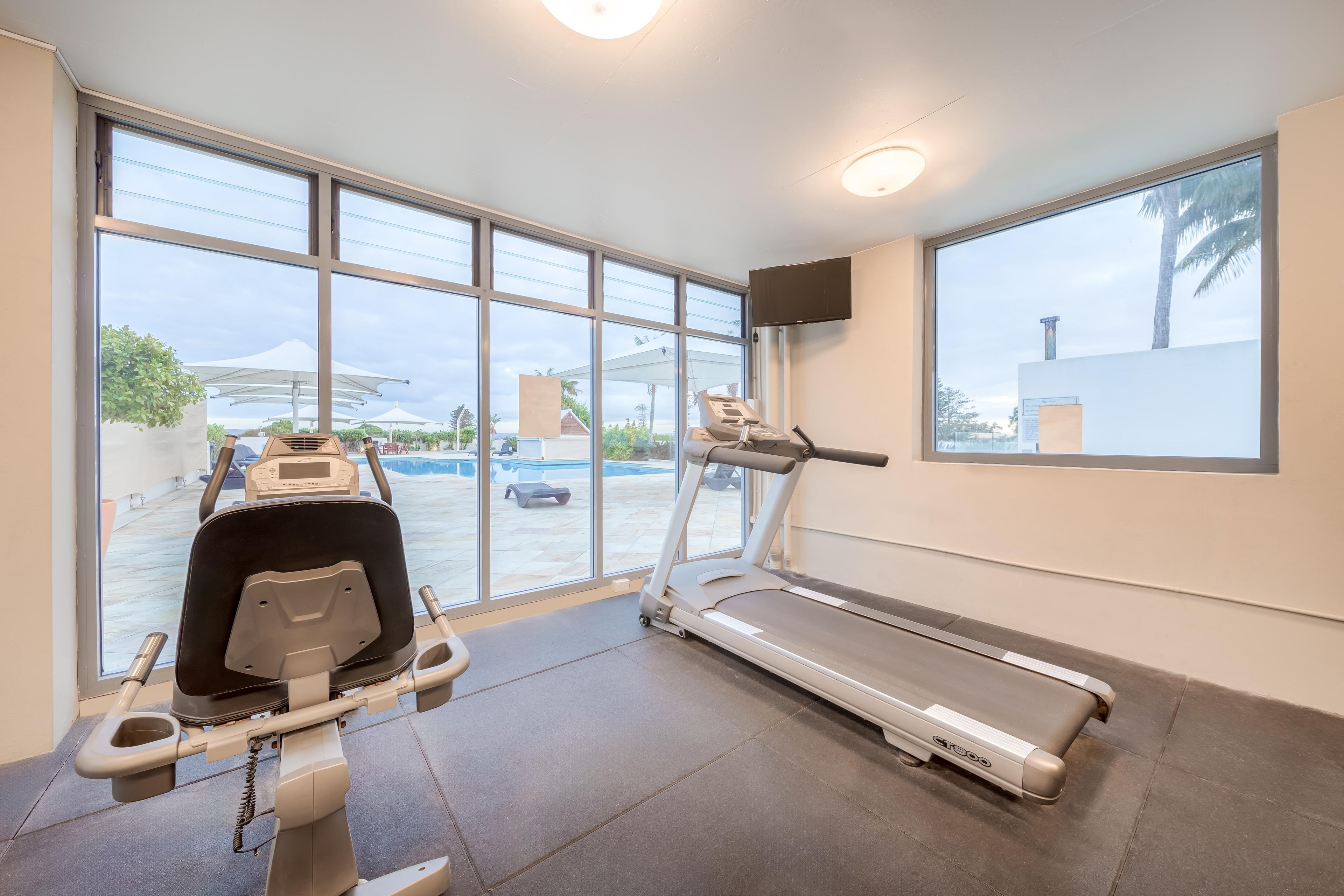 A gym with a treadmill and a stationary bike, large windows with a view of a pool and palm trees.
