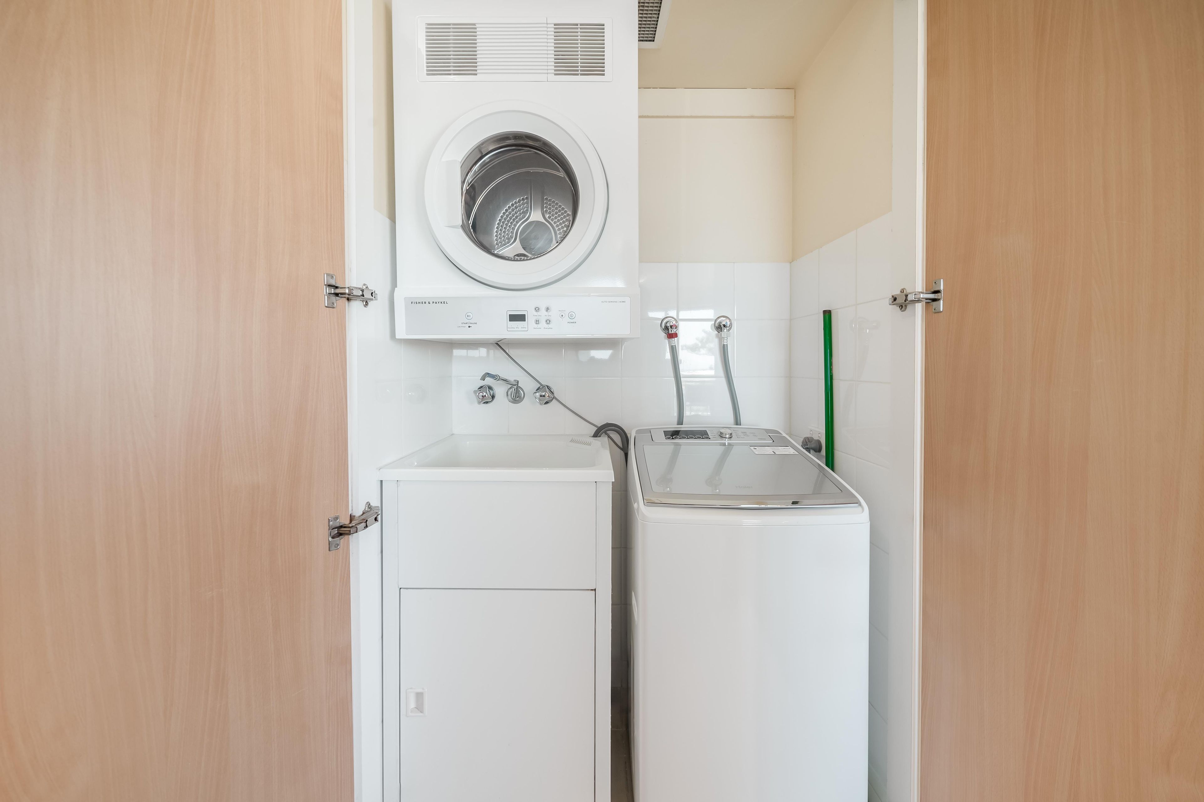 A compact laundry area with a front-loading washer and dryer, a small sink, and a green broom.