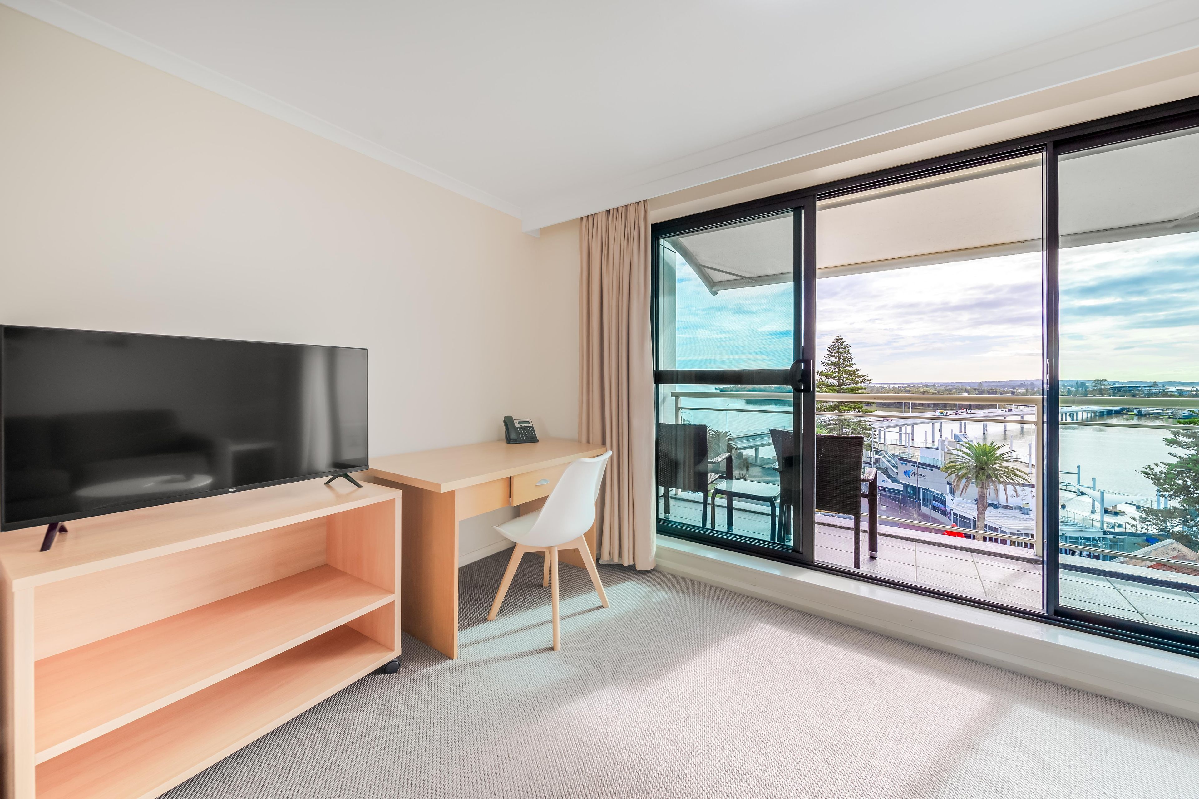 A hotel room with a large window, a flat-screen TV, a desk, and a chair.
