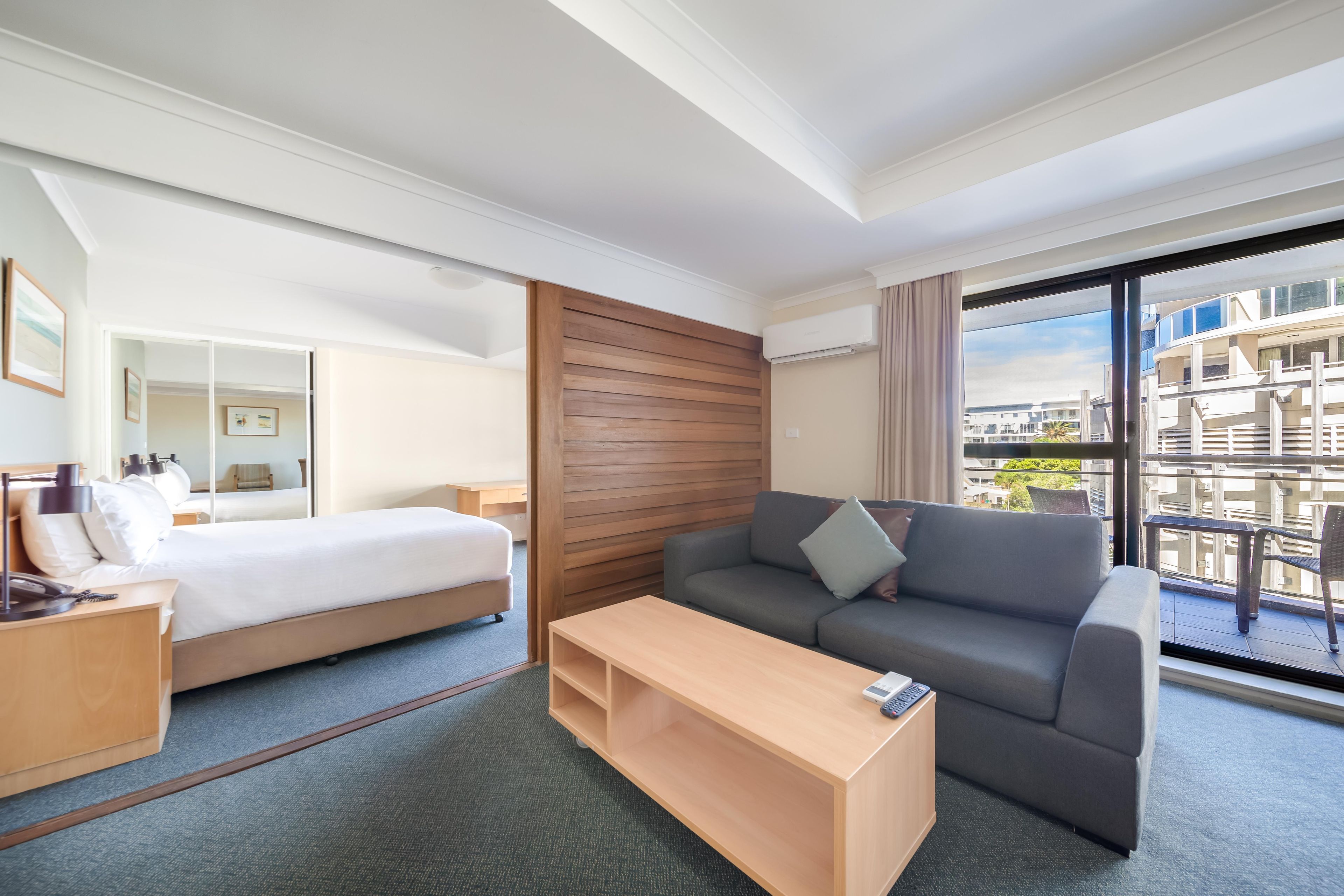 Ein Hotelzimmer mit einem Bett, einem Schlafsofa, einem Holztischchen und einem großen Fenster mit Blick auf Gebäude.