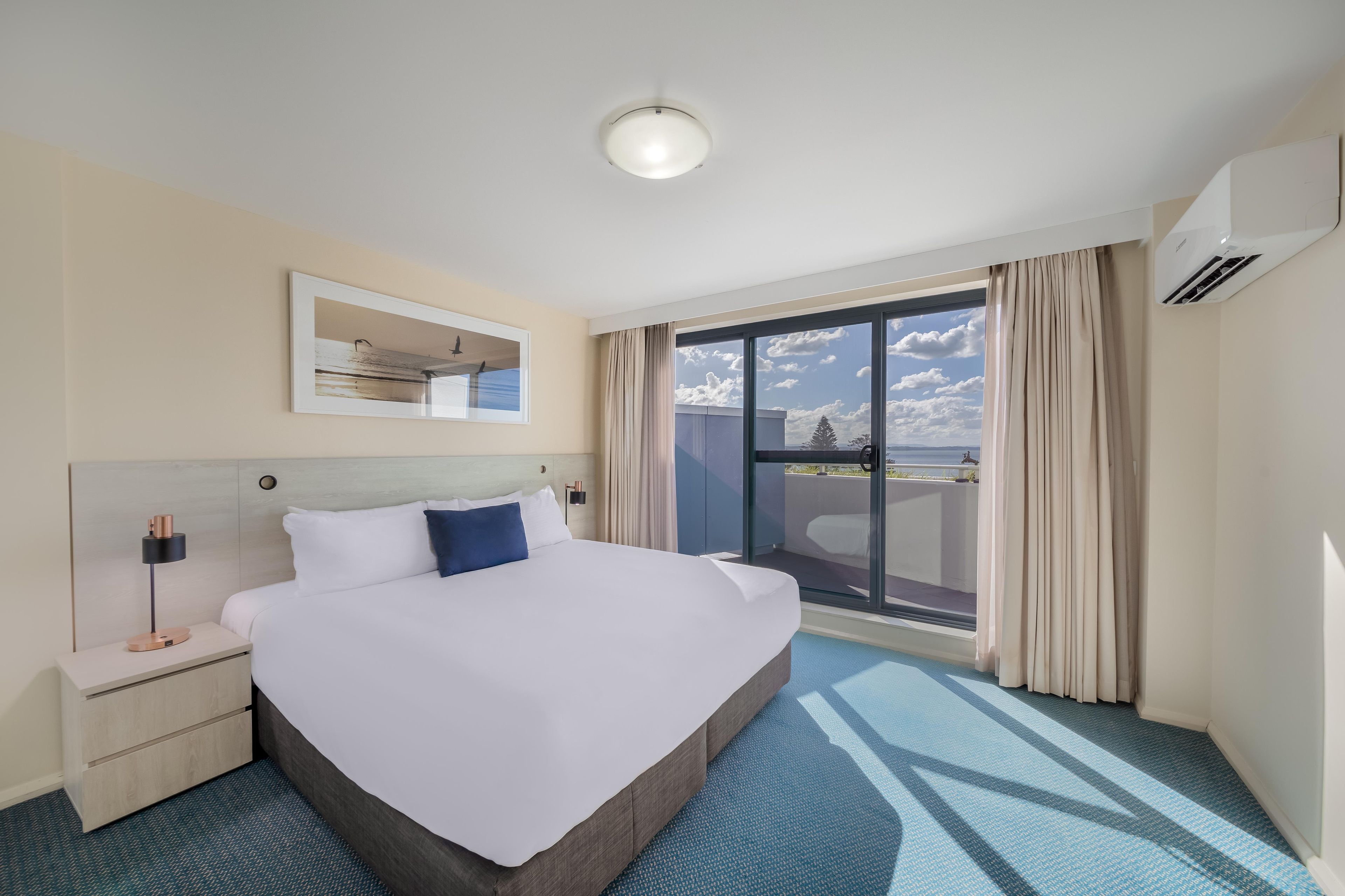 Habitación de hotel con una cama grande, una mesita de noche, una lámpara, una luz en el techo, una ventana con cortinas y un equipo de aire acondicionado.