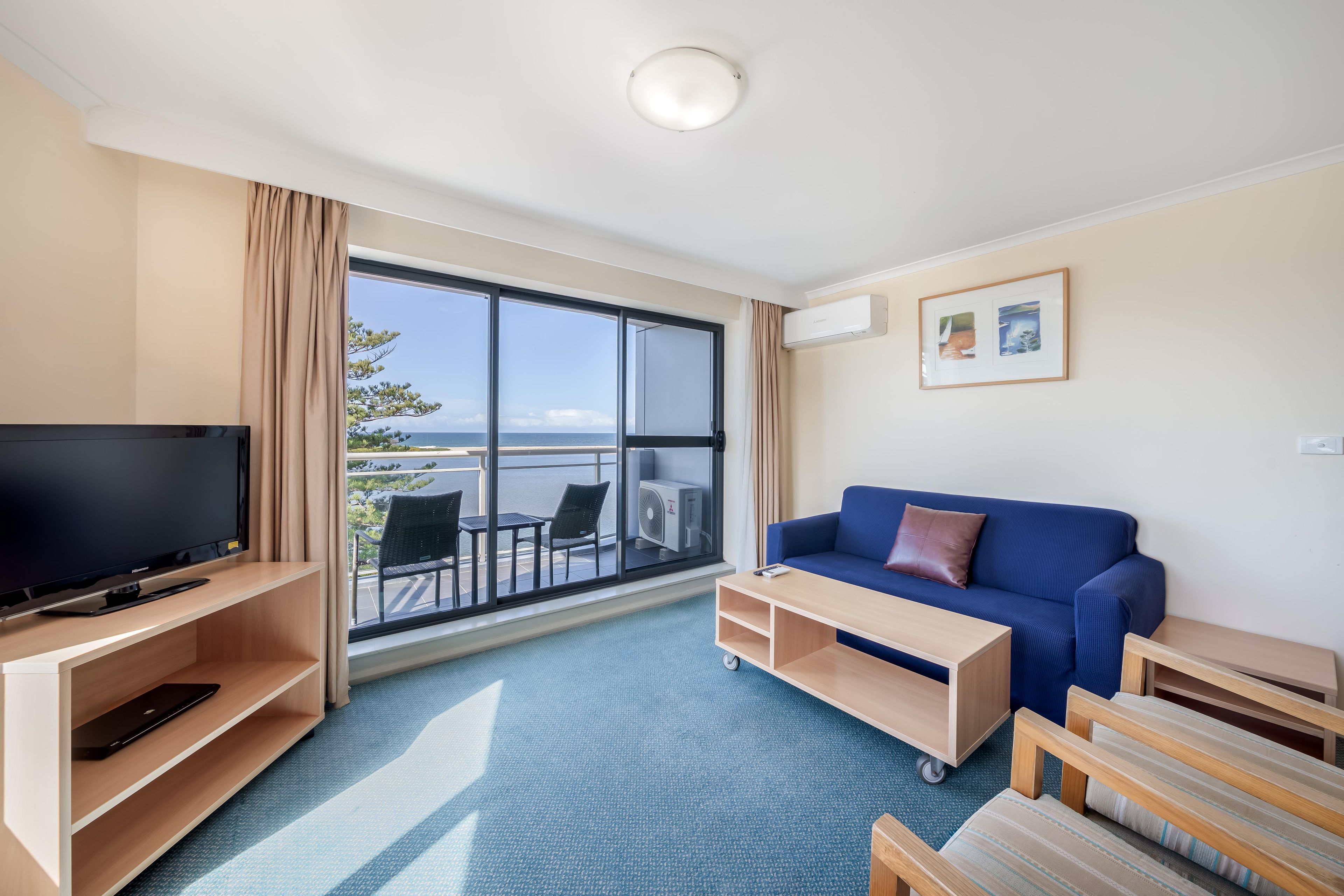 A hotel room with a large window, a flat-screen TV, a blue sofa, and a small wooden table.