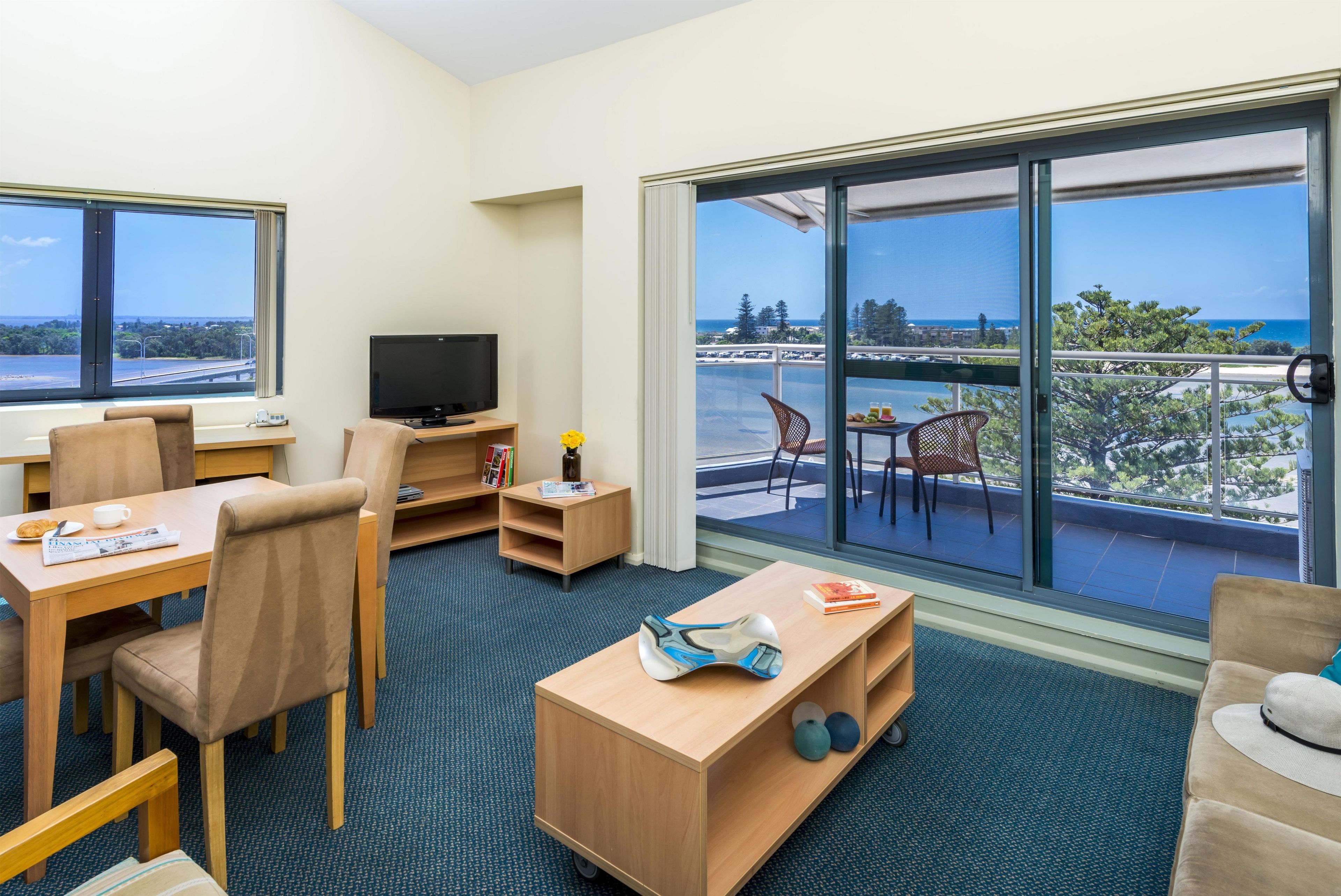 A hotel room with a balcony, a TV, a dining area, and a view of the ocean.