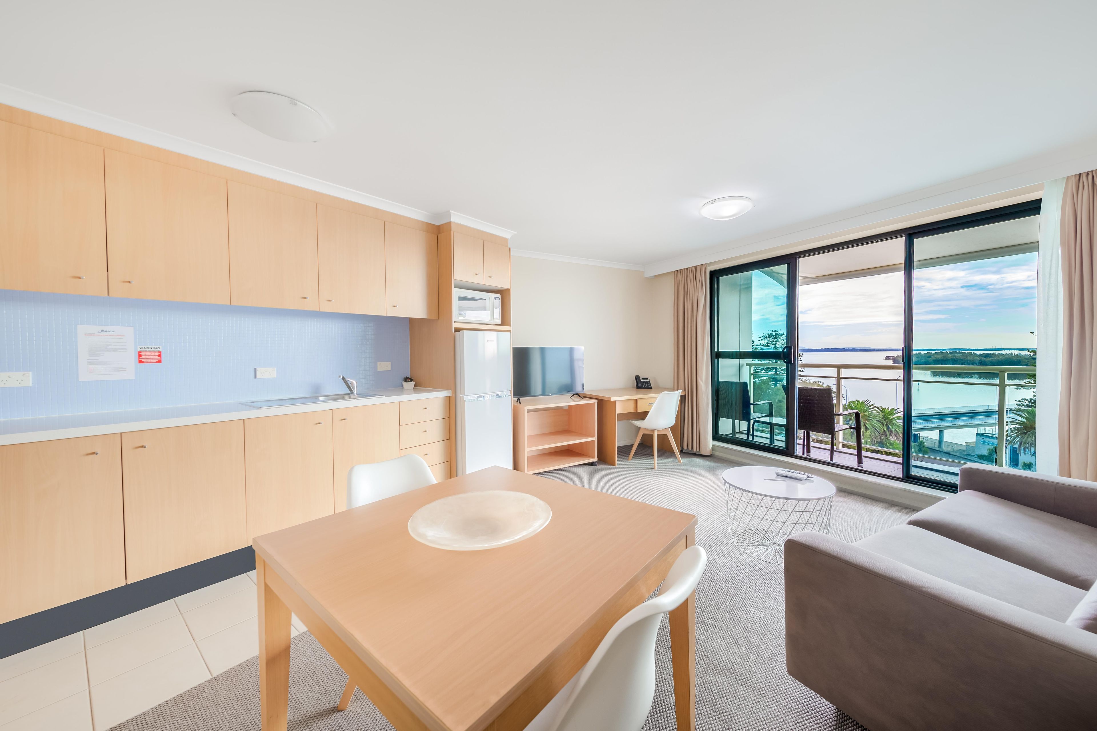 A modern kitchen with wooden cabinets, a microwave, and a refrigerator. There is a dining table with chairs, a sofa, and a large window offering a view of the city.