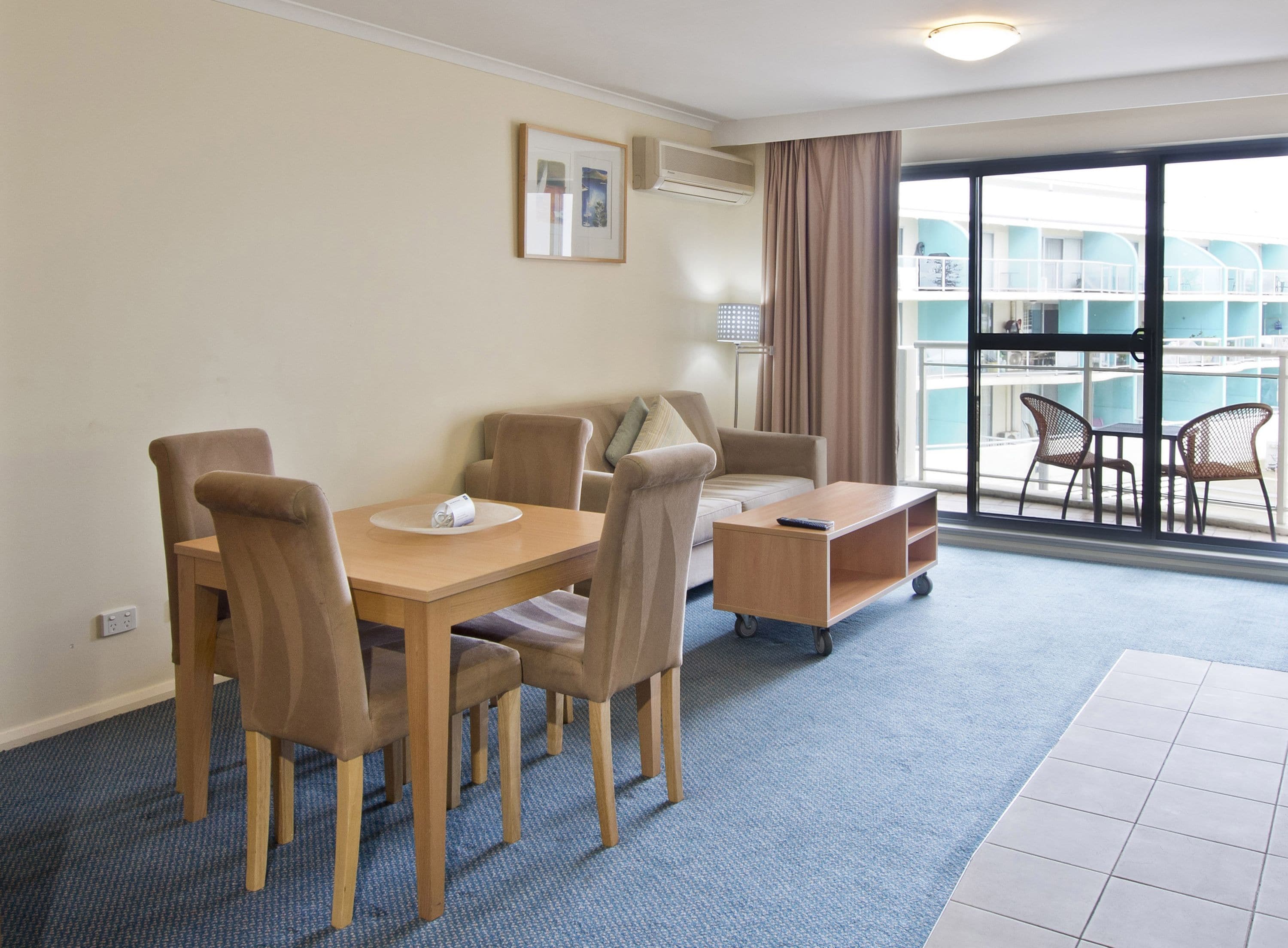 Habitación de hotel con una mesa de comedor, sillas, un pequeño sofá, una mesita auxiliar y vistas a una piscina y un edificio a través de amplios ventanales.
