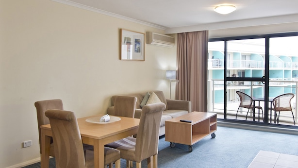 Ein Hotelzimmer mit Esstisch, Stühlen, einer kleinen Couch, einem Beistelltisch und Blick auf ein Schwimmbad und ein Gebäude durch große Fenster.