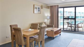 Habitación de hotel con una mesa de comedor, sillas, un pequeño sofá, una mesita auxiliar y vistas a una piscina y un edificio a través de amplios ventanales.