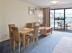 A hotel room with a dining table, chairs, a small sofa, a side table, and a view of a pool and building through large windows.